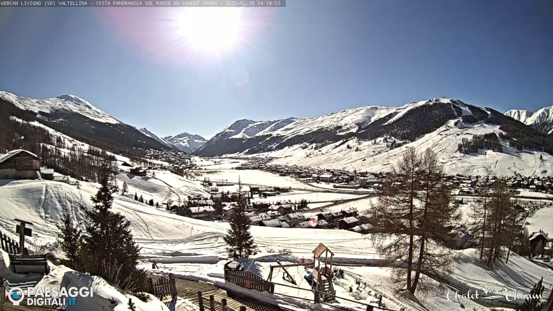 A sunny, snow-covered mountain valley village is visible from a high vantage point, with several evergreen trees in the foreground.