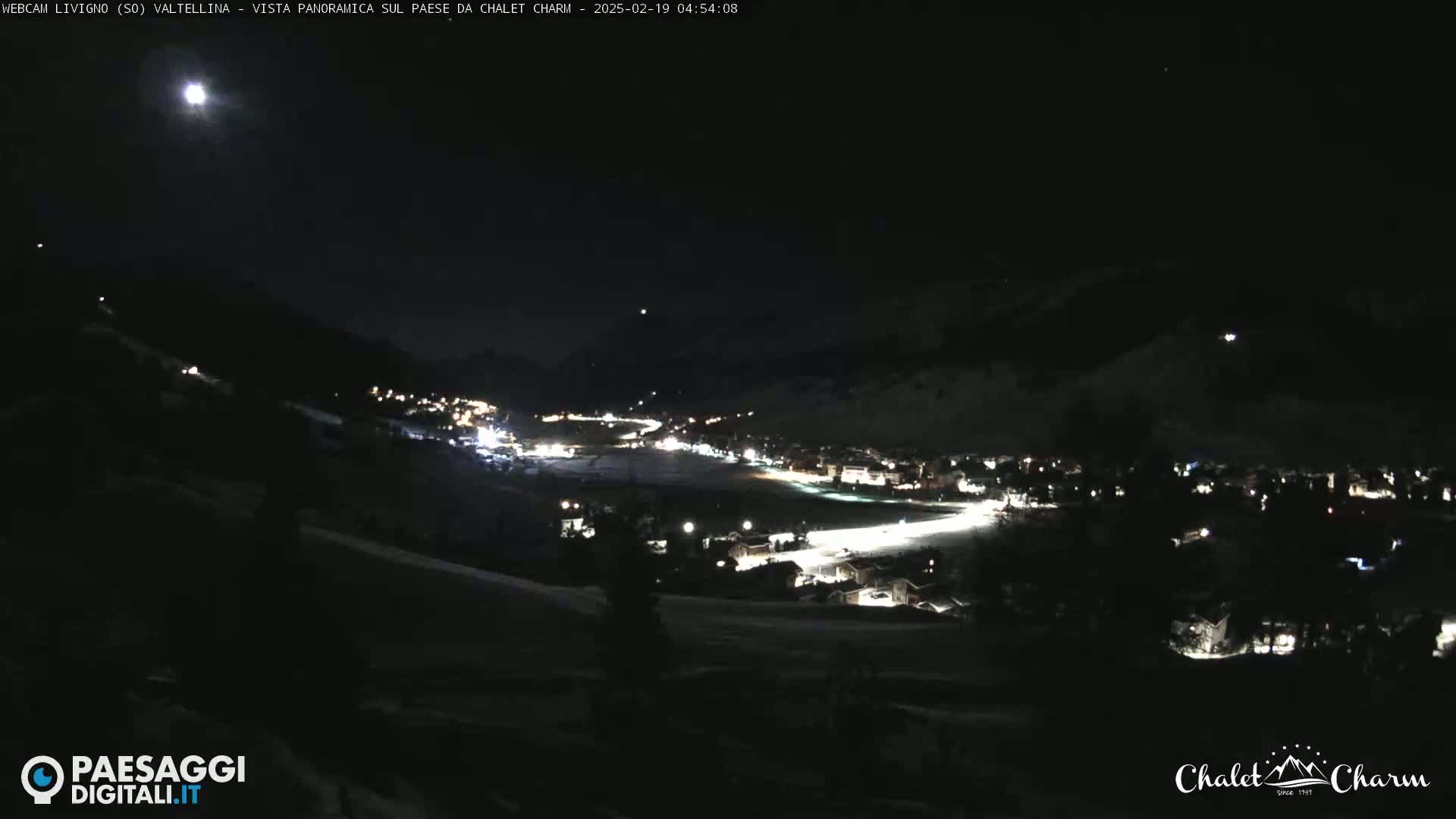A nighttime, panoramic view of a snow-covered town illuminated by lights under a bright moon.