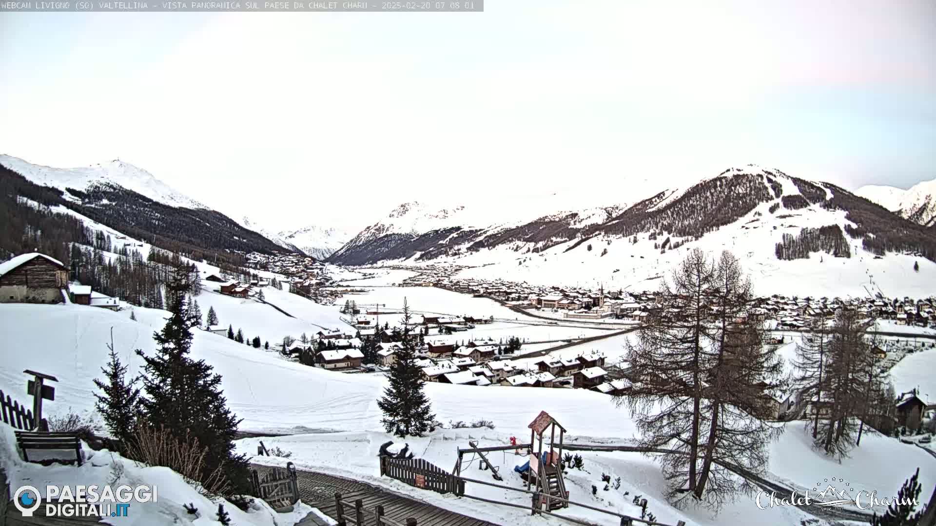 A snow-covered village nestled in a valley between snow-capped mountains under a cloudy sky.
