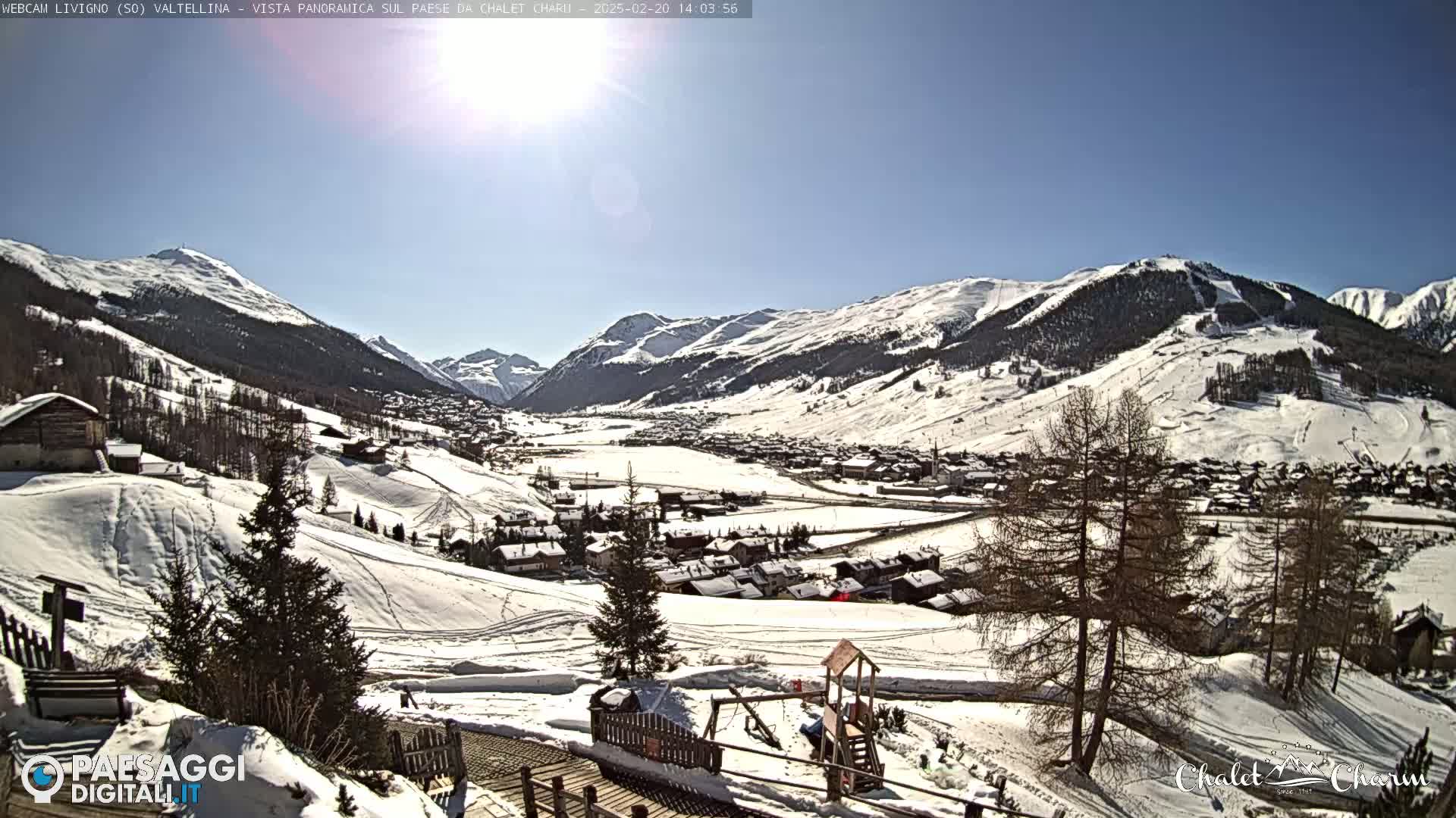 A sunny, snow-covered mountain valley village scene, with a children's playground in the foreground.