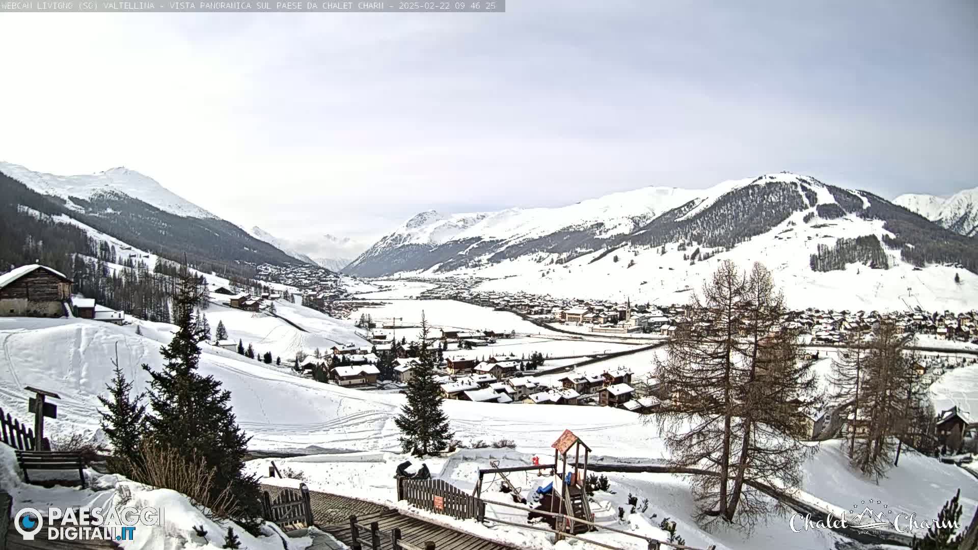 A snow-covered village nestled in a valley between snow-capped mountains under a mostly cloudy sky.