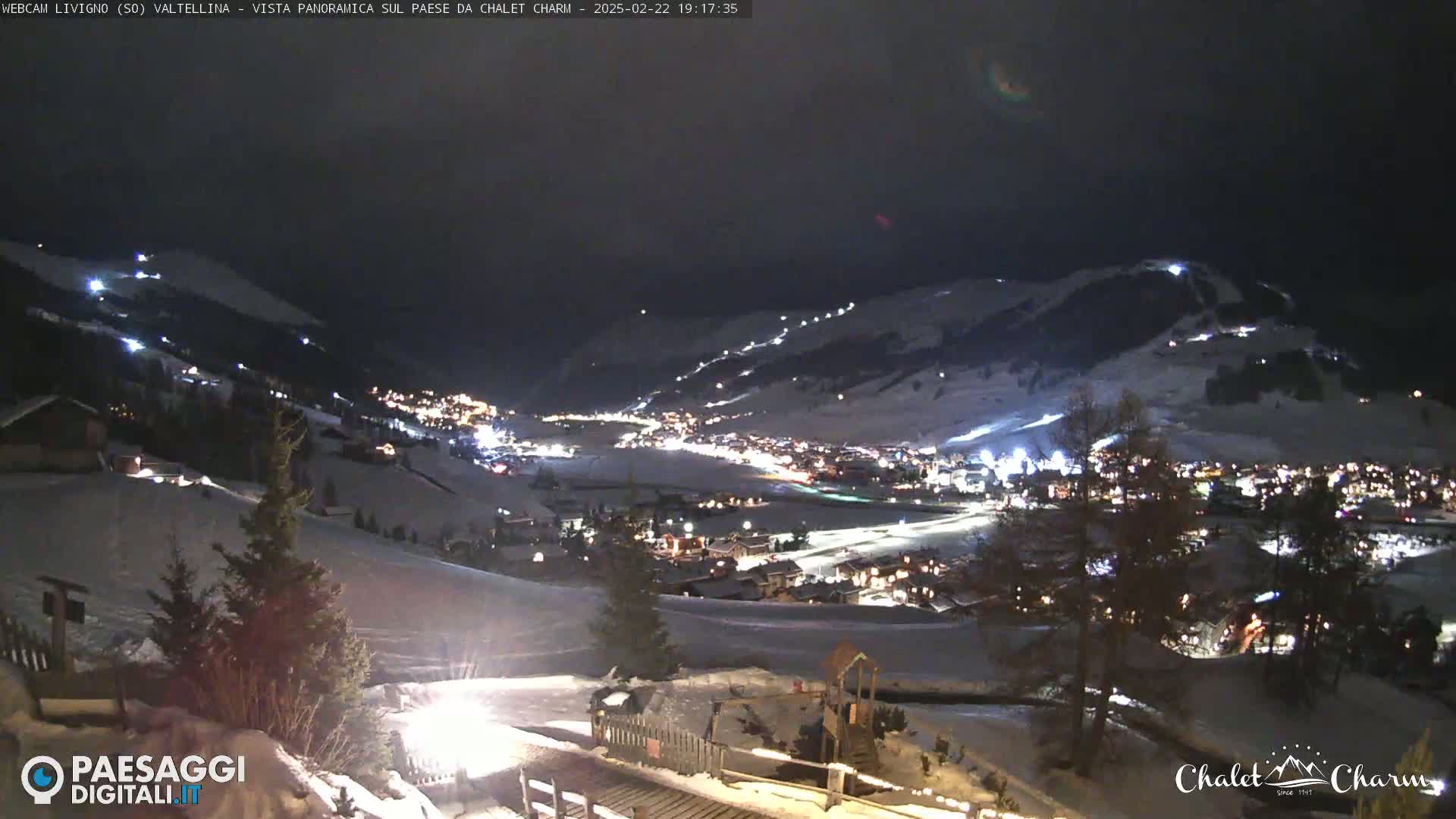 A nighttime, panoramic view of a snow-covered town nestled in a valley, illuminated by streetlights and lights on a nearby ski slope.