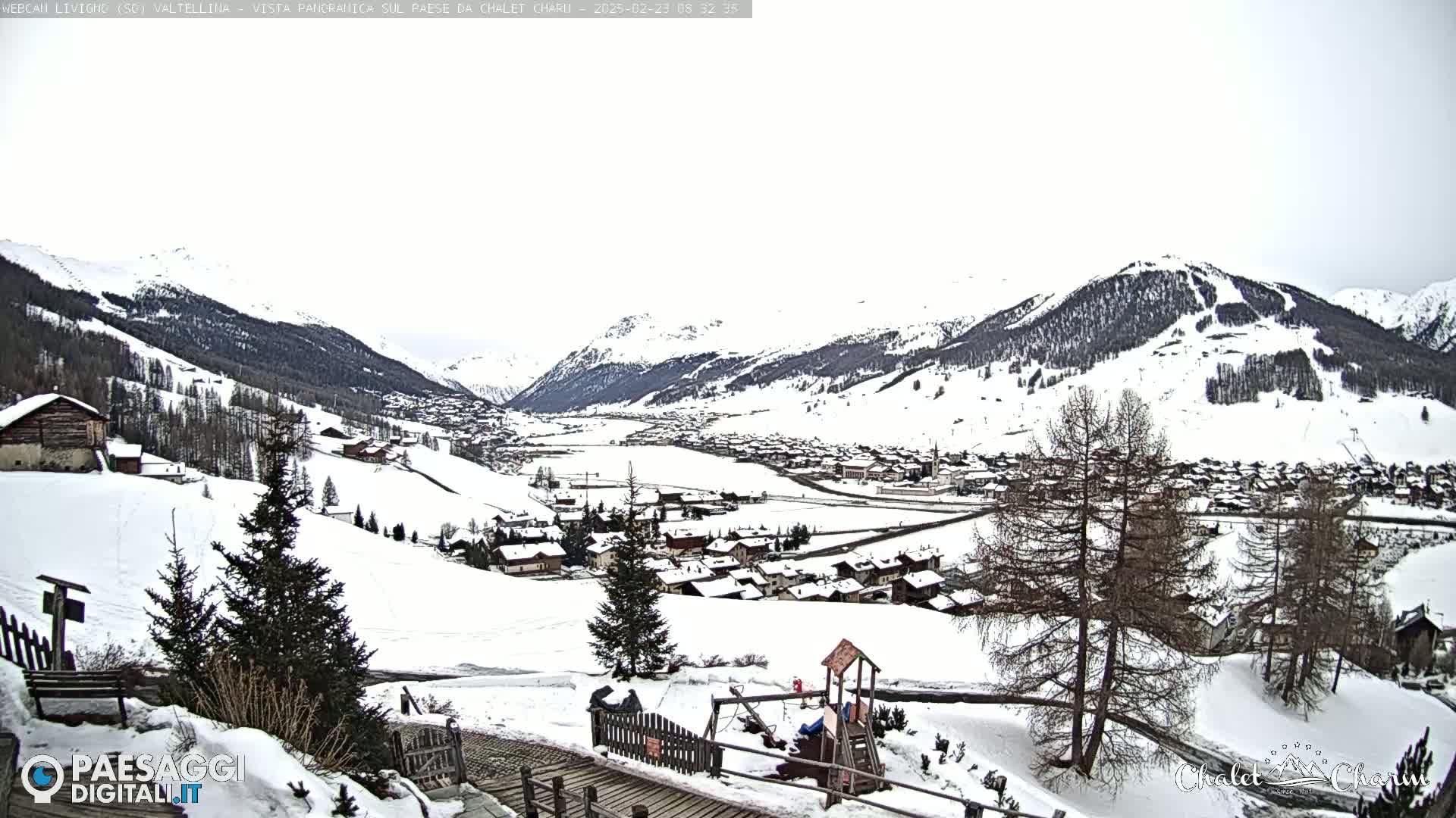 A snow-covered mountain valley village is viewed from a hillside, with a children's playground in the foreground under an overcast sky.