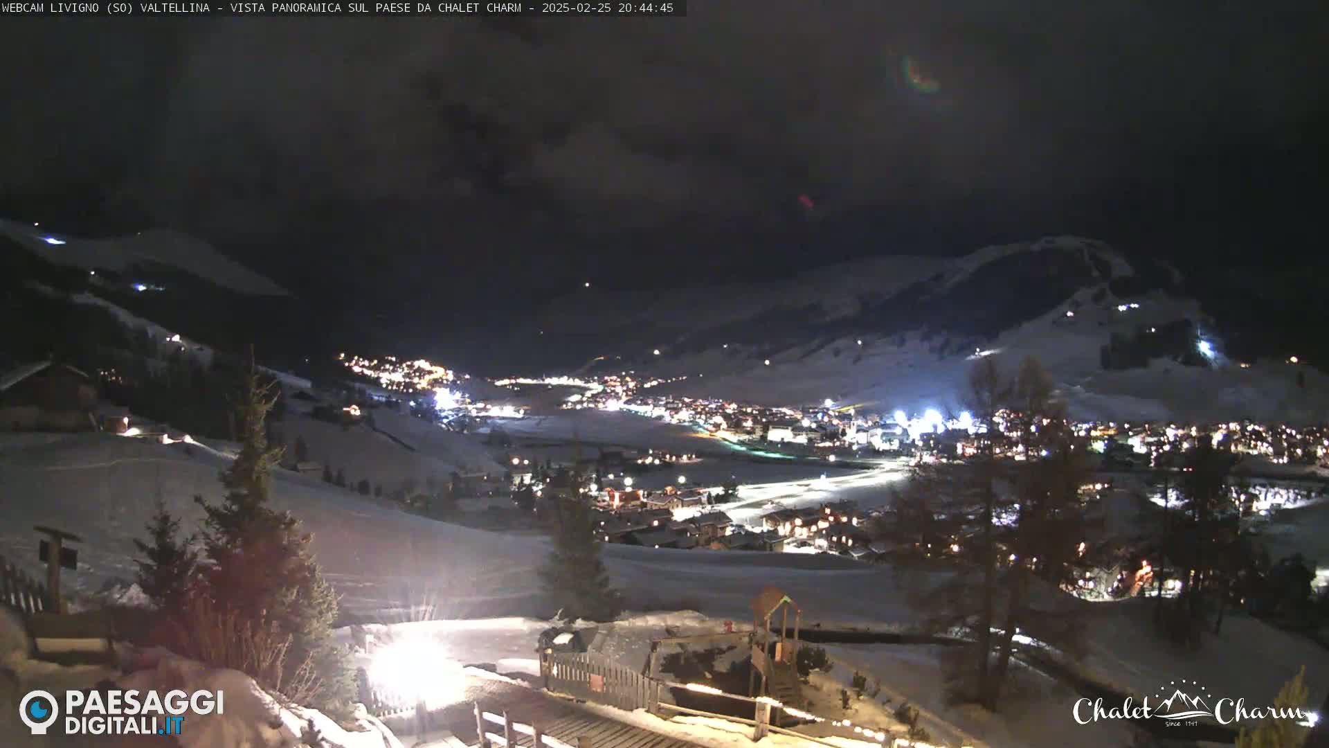 A nighttime, panoramic view of a snow-covered town illuminated by lights, under a dark, cloudy sky.