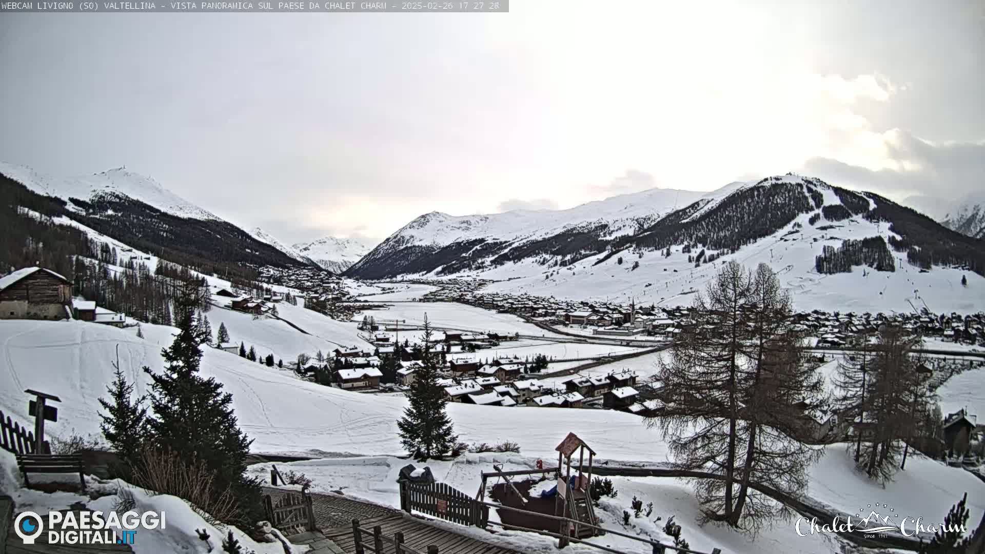 A snow-covered village nestled in a valley between snow-capped mountains under a cloudy sky.