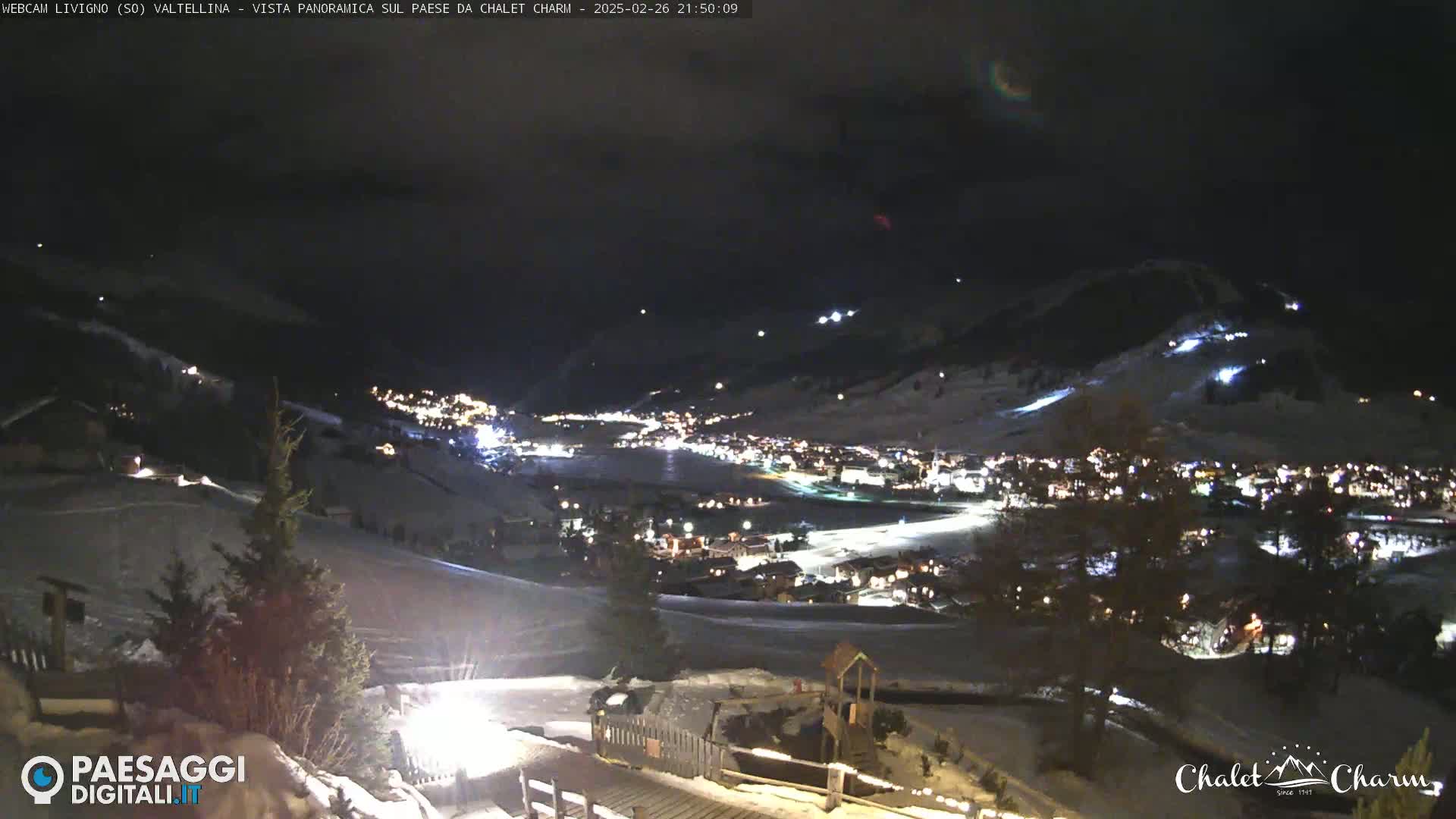 A nighttime, panoramic view of a snow-covered town nestled in a valley, illuminated by lights, under a dark sky with a few faint clouds.