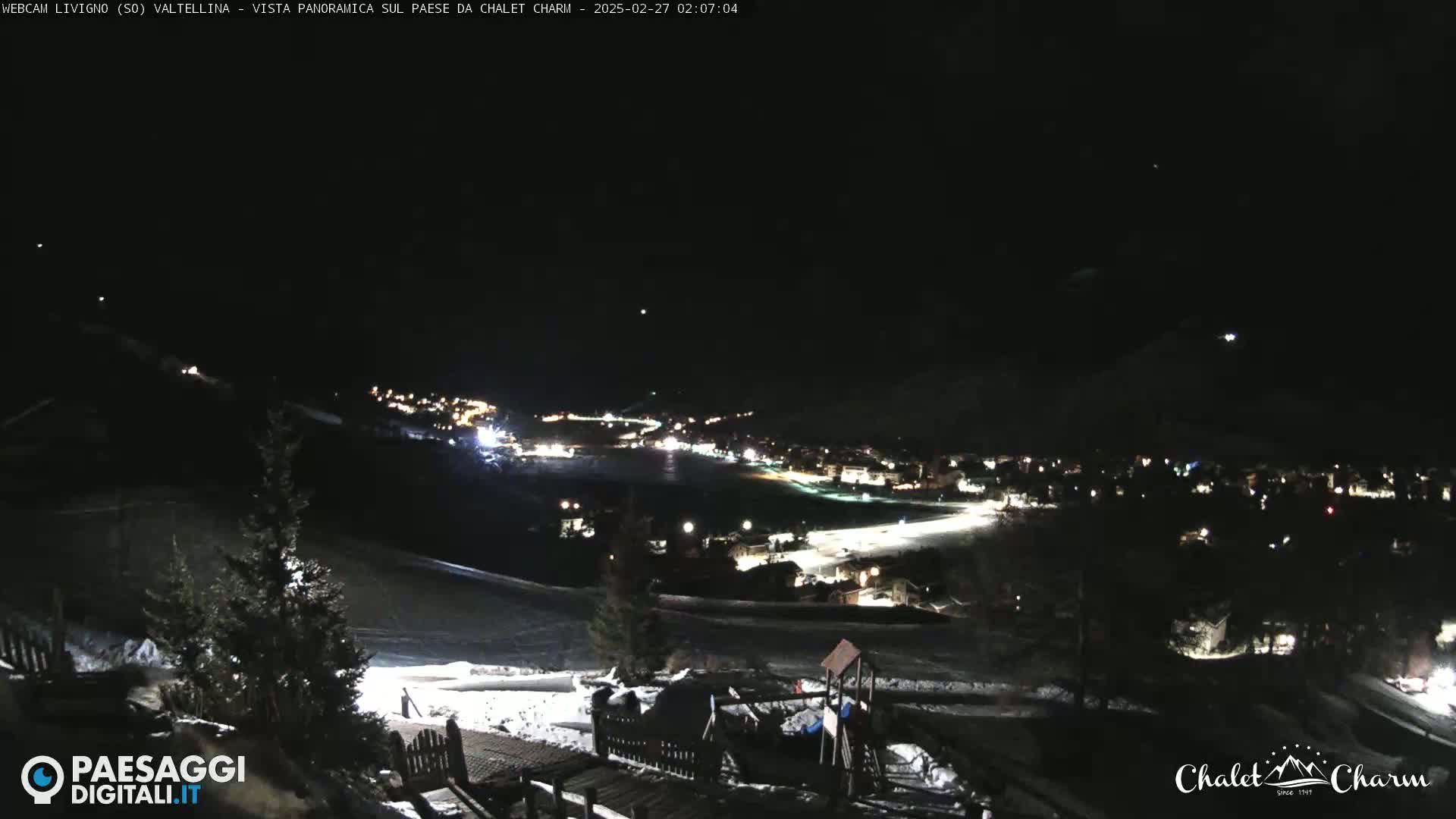 A nighttime, panoramic view of a snow-covered village with illuminated buildings and a playground, under a clear, dark sky.
