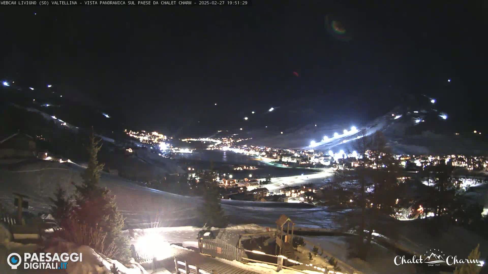 A nighttime, panoramic view of a snow-covered town nestled in a valley, illuminated by lights, with ski slopes visible on the surrounding hills.