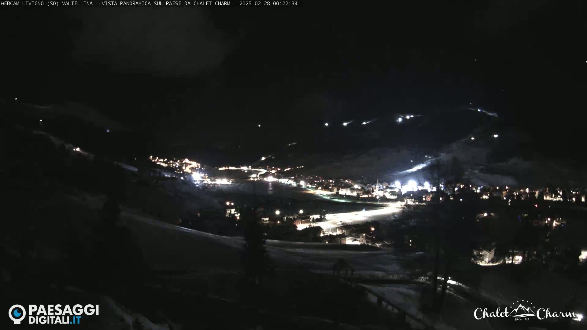 A nighttime, panoramic view of a snow-covered town illuminated by streetlights and building lights, with several bright lights visible on the surrounding hills.