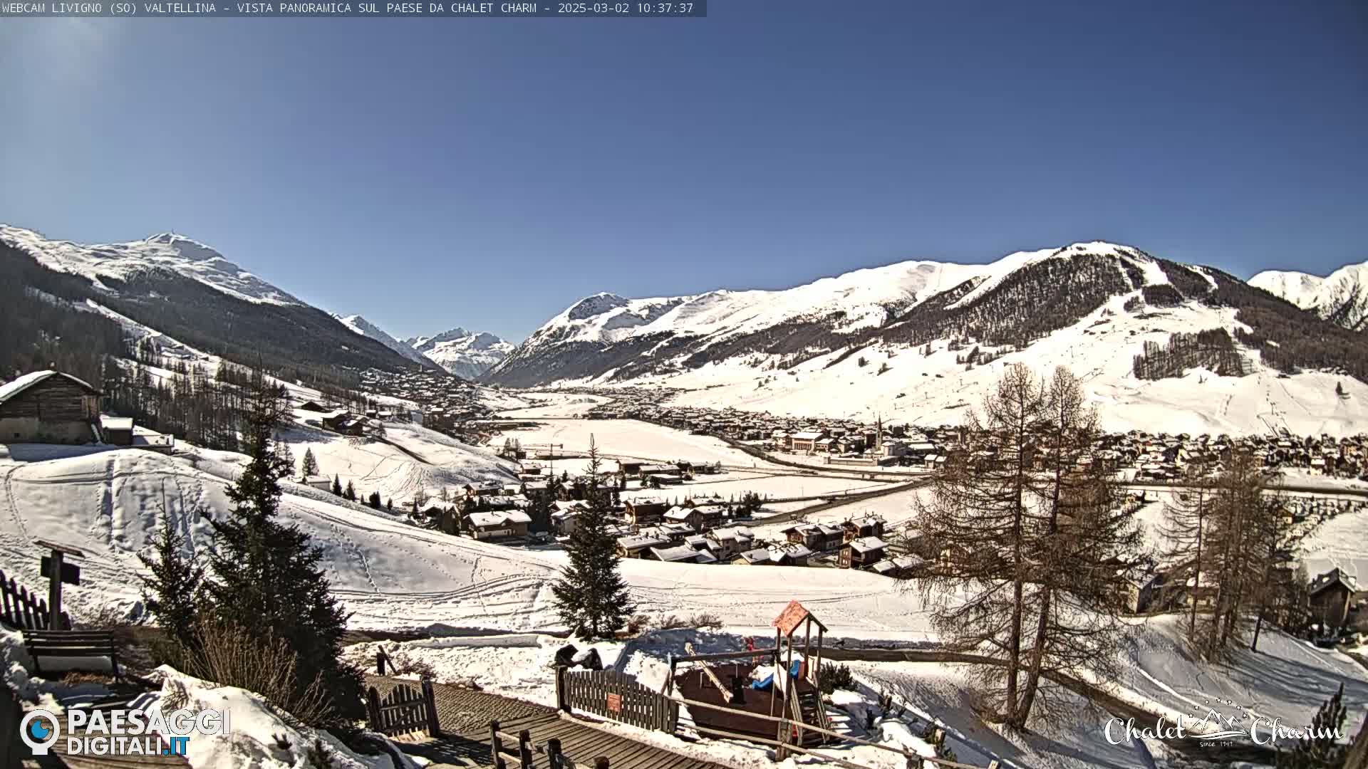 A snow-covered mountain village is visible from a hilltop on a sunny day.