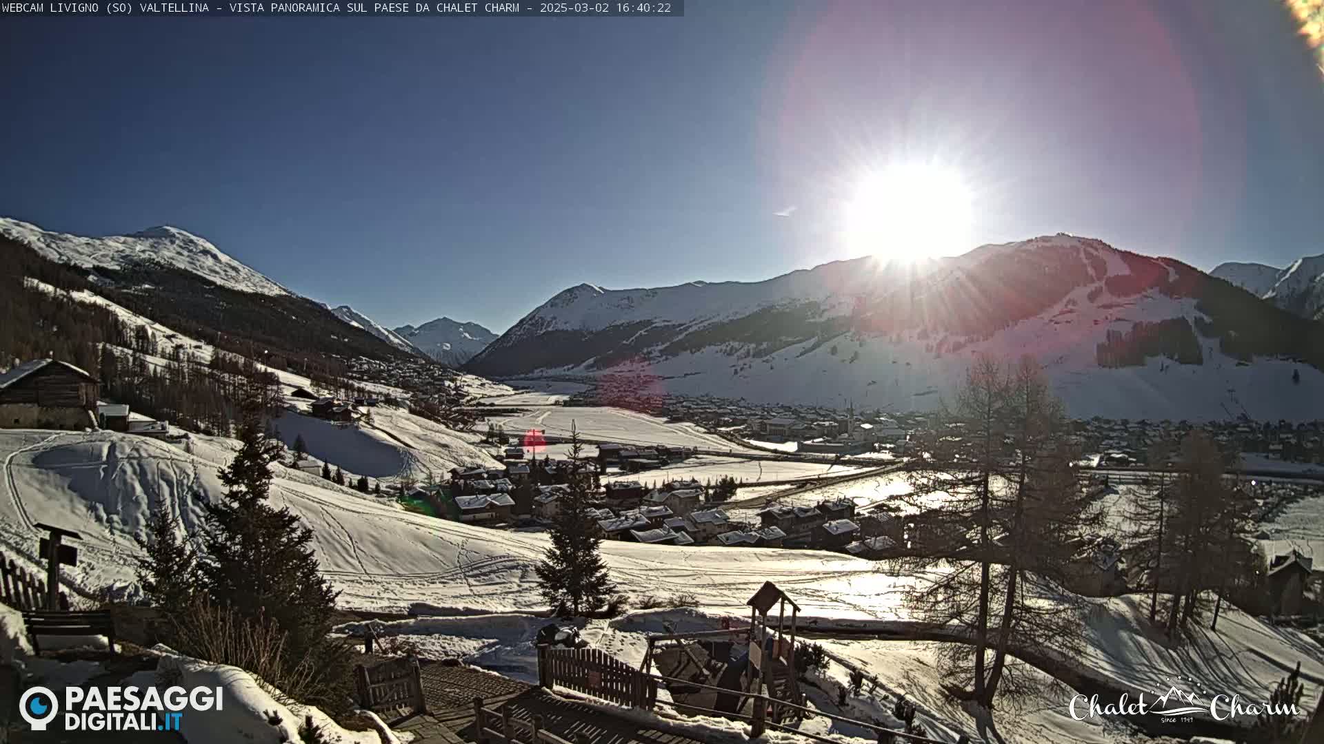 A sunny, snow-covered mountain valley town is viewed from a hilltop, showcasing a playground and surrounding snowy landscape.