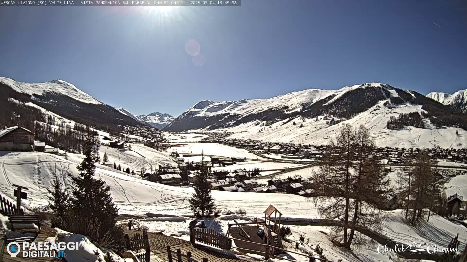 A sunny, snow-covered mountain valley village is viewed from a hillside, with a children's playground visible in the foreground.
