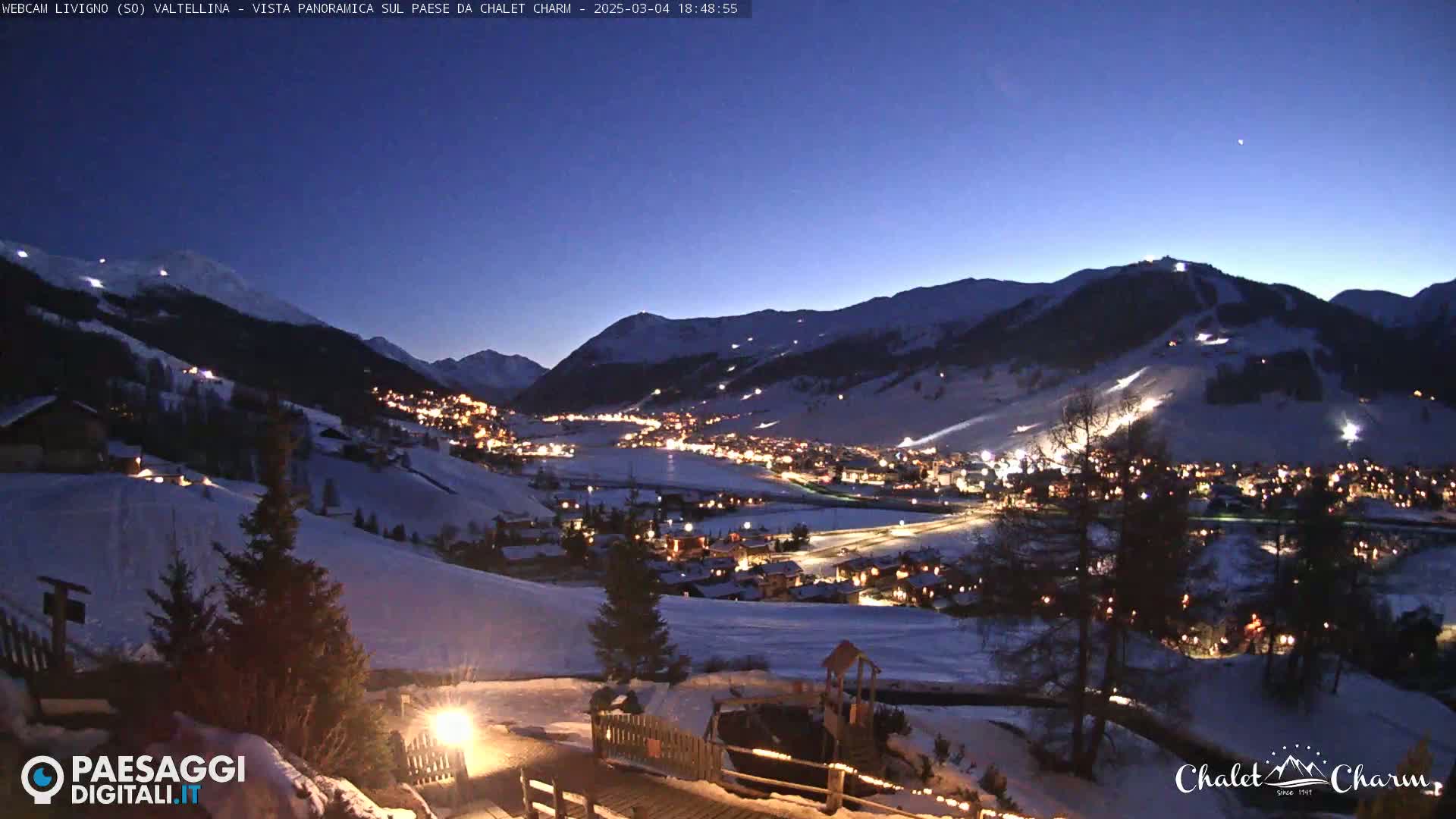 A nighttime view of a snow-covered village nestled in a valley between illuminated mountains under a twilight sky.