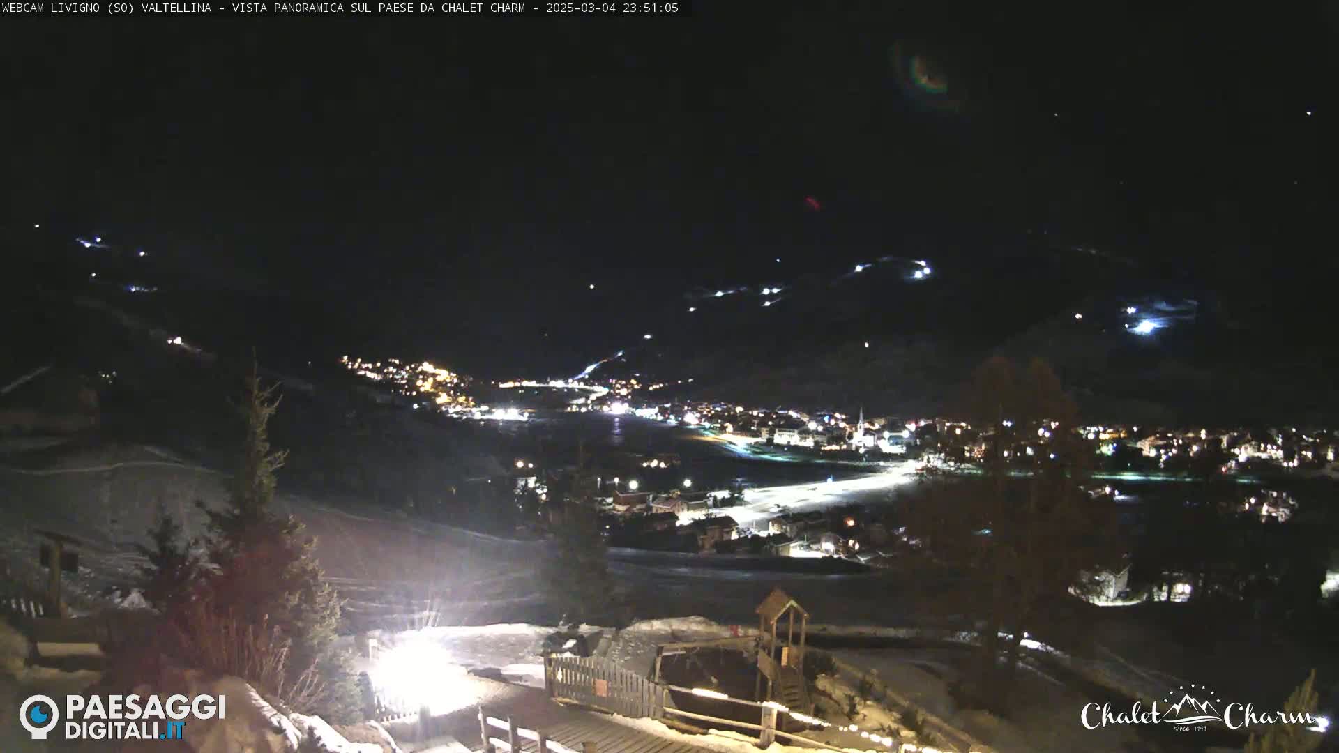 A nighttime, panoramic view of a snow-covered town illuminated by lights, with a clear sky above and faintly visible lights in the distance, possibly from skiers or other activities.