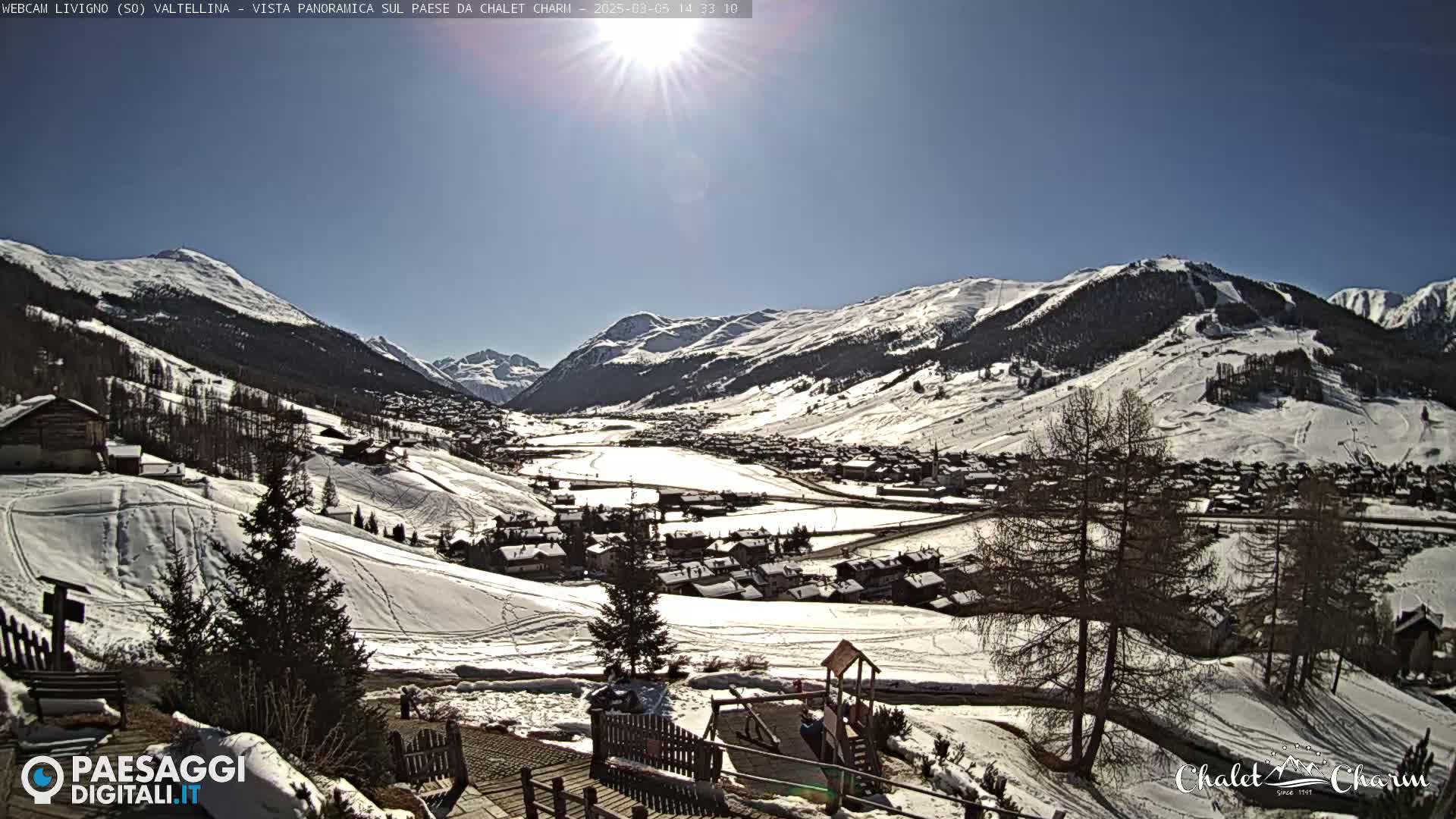 A sunny, snow-covered mountain valley village is viewed from a high vantage point, with several trees and a playground visible in the foreground.