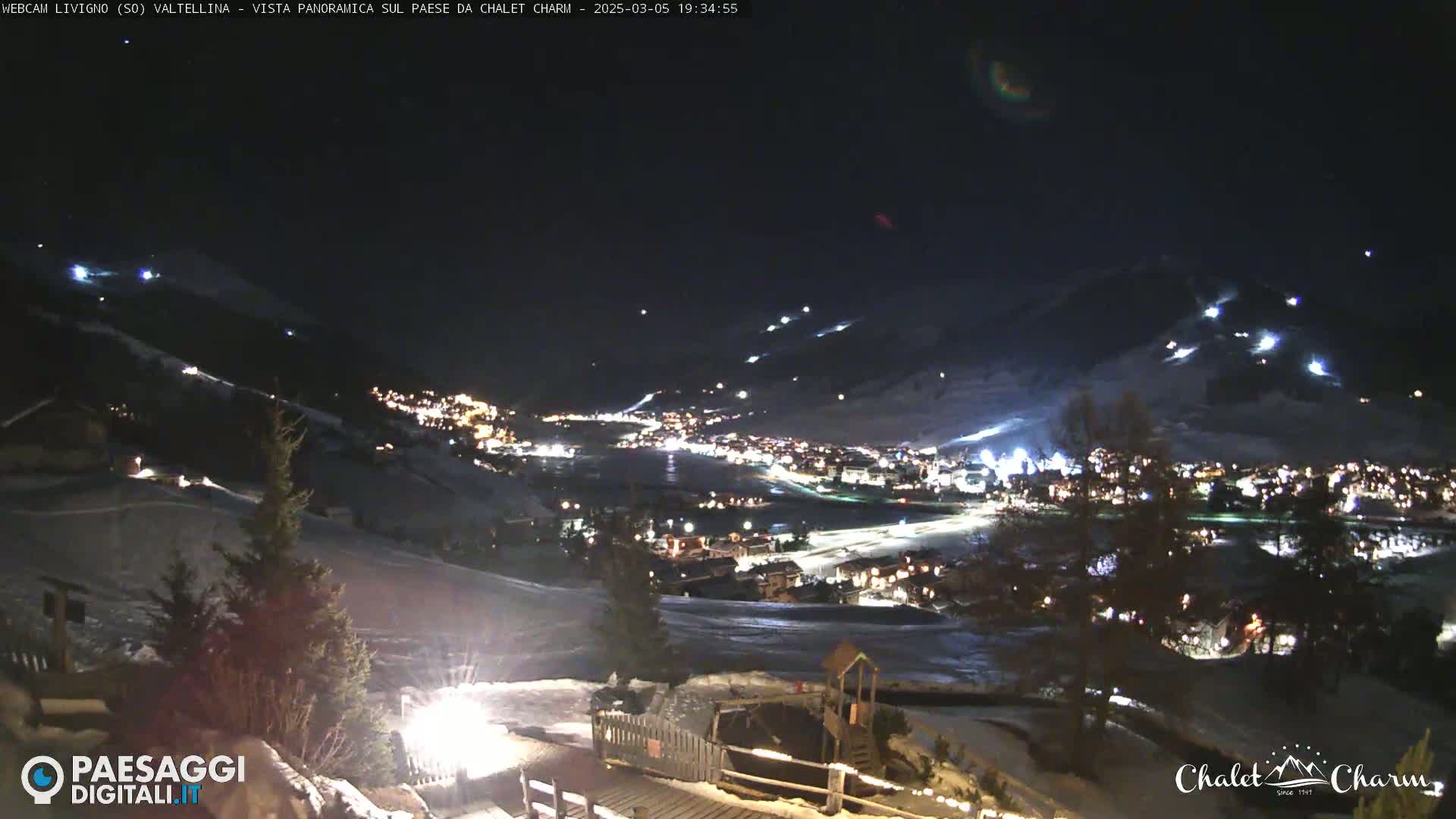 A nighttime, panoramic view of a snow-covered town illuminated by lights, with sparsely lit ski slopes visible on surrounding hills under a dark, clear sky.