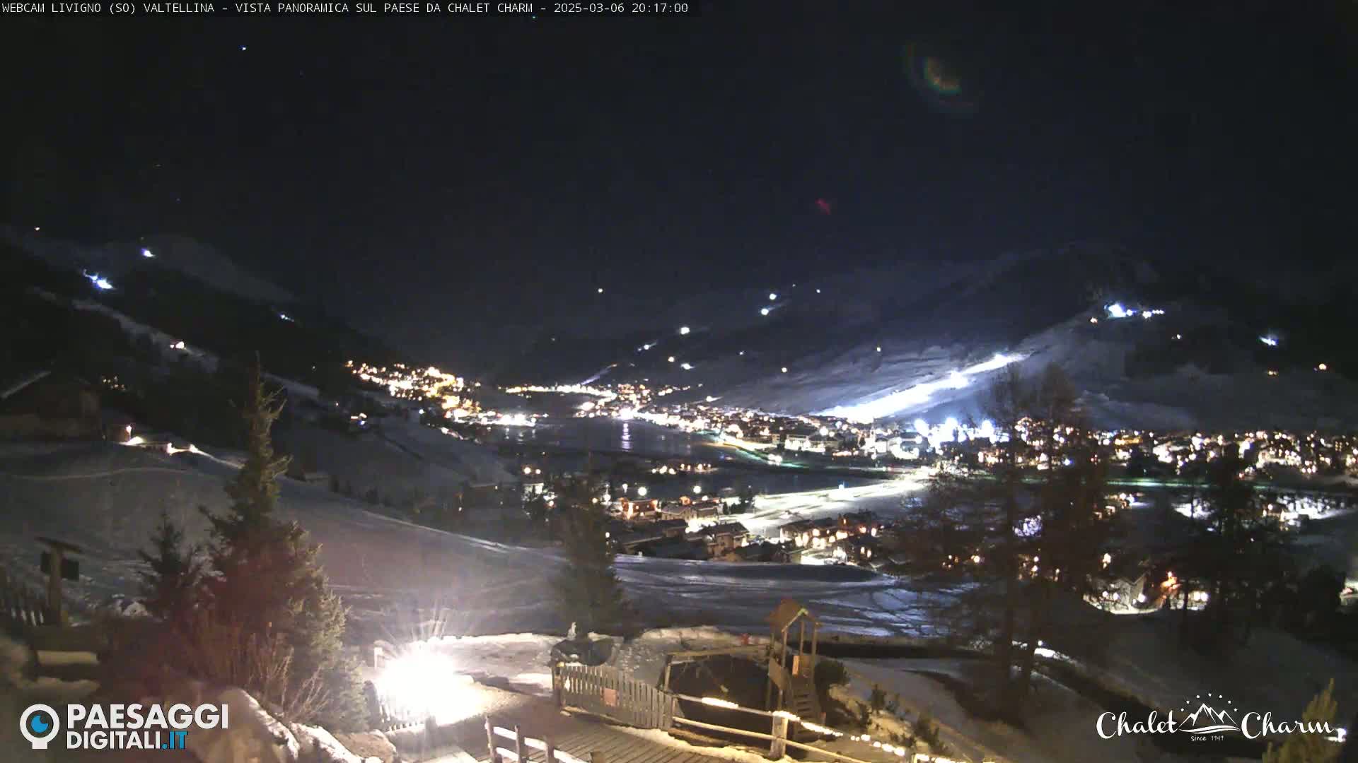 A nighttime, panoramic view of a snow-covered village illuminated by lights, nestled in a valley under a clear, dark sky.