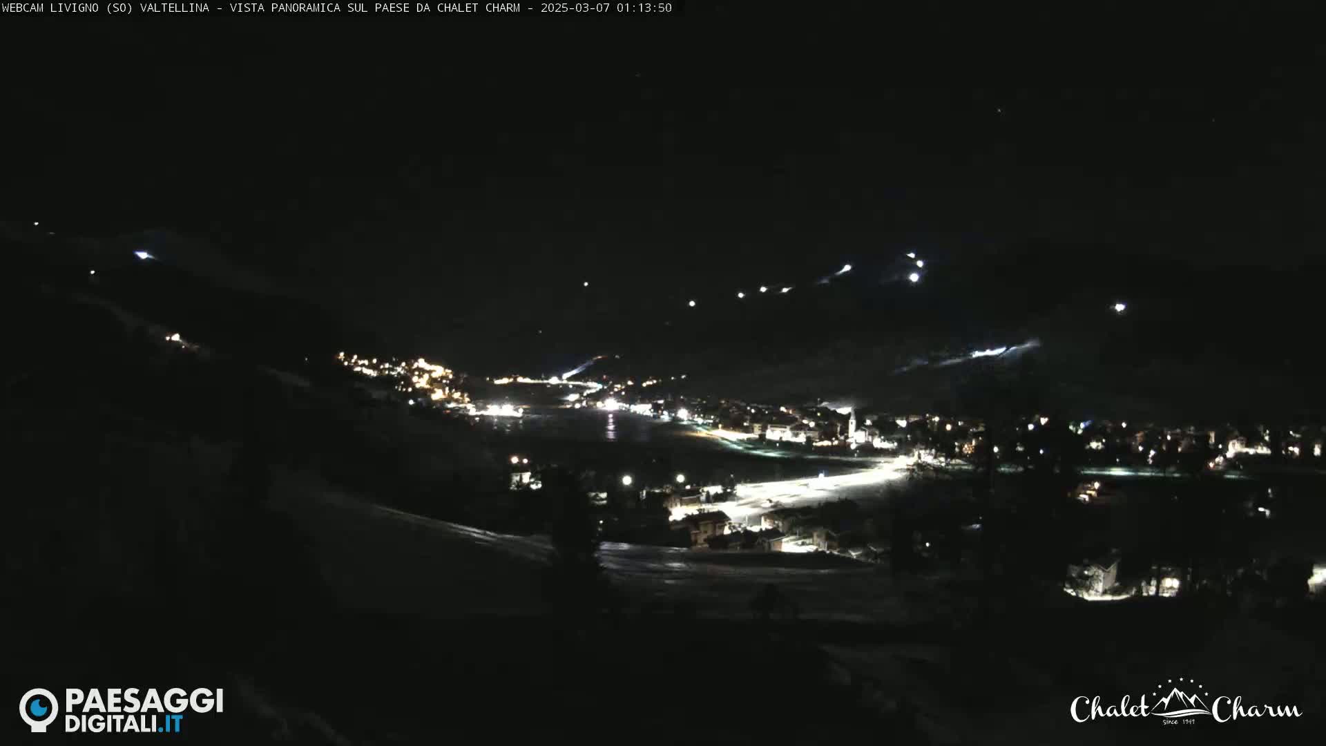A nighttime, panoramic view of a snow-covered town illuminated by lights, with faint light trails suggesting movement in the snowy hills above.