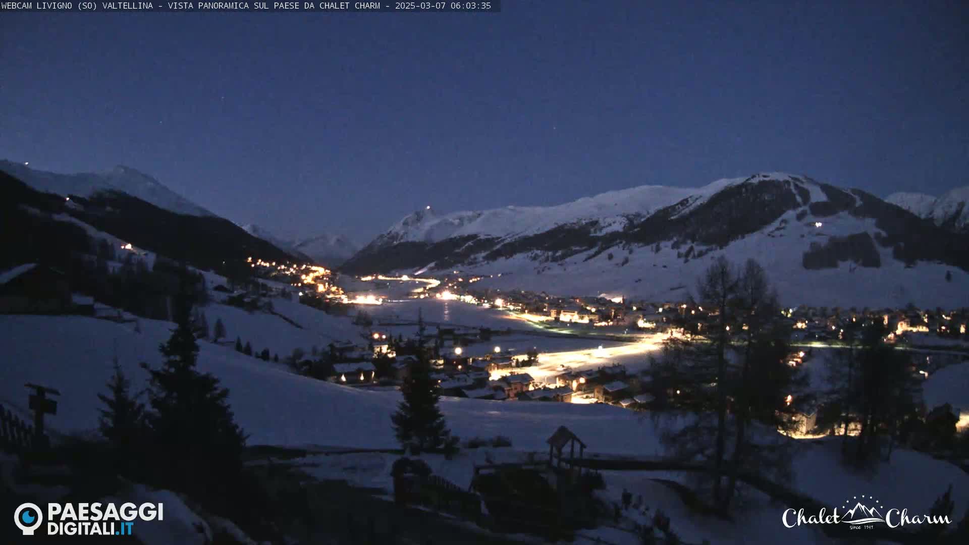 A snow-covered mountain village is illuminated at night under a clear, dark blue sky.