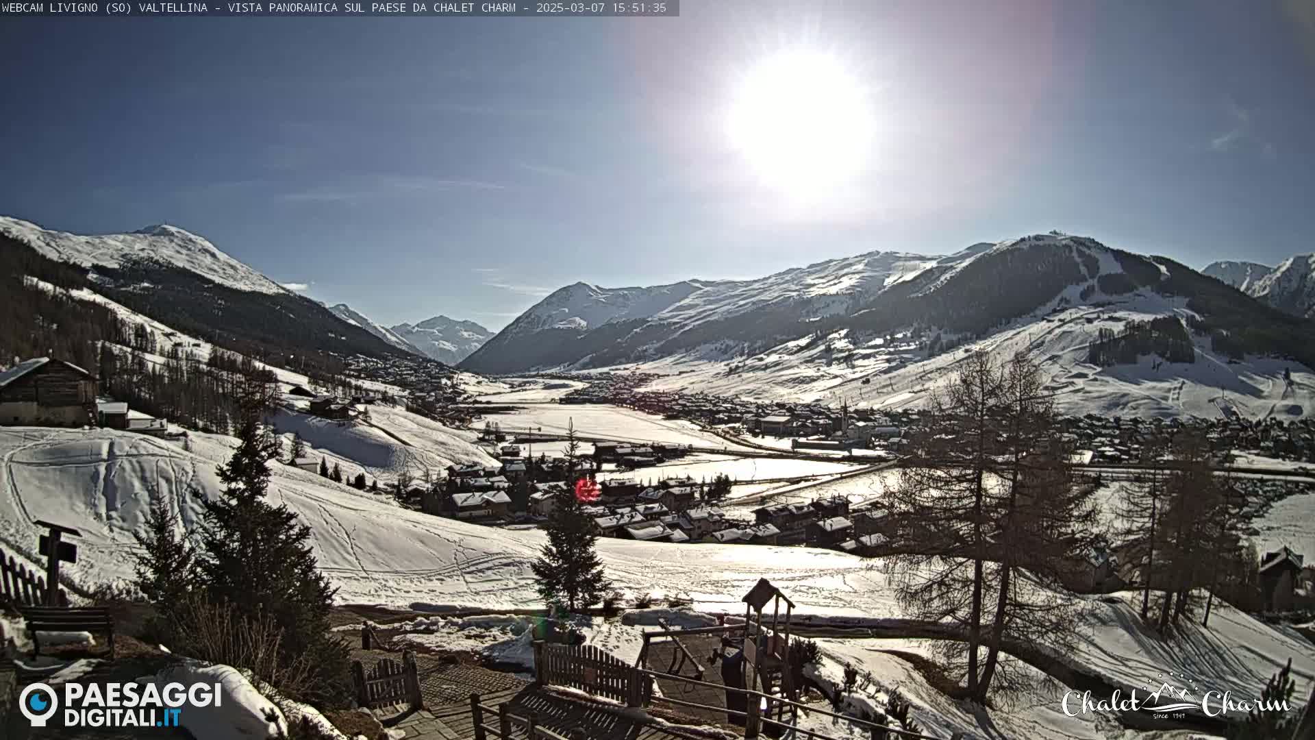 A sunny, snow-covered mountain valley town is viewed from a hillside, with a playground visible in the foreground.