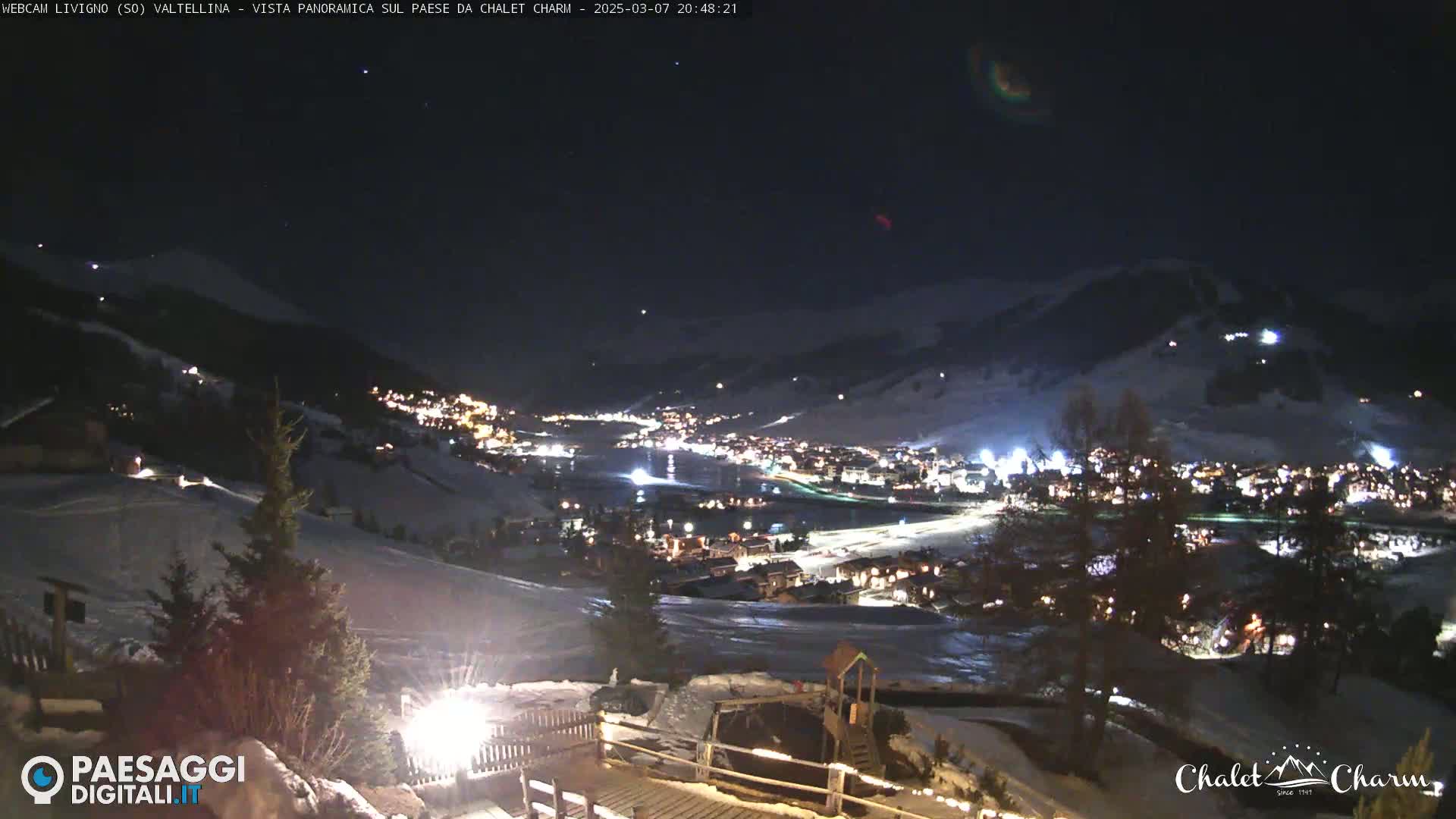 A nighttime, panoramic view of a snow-covered town illuminated by lights, nestled in a valley between mountains under a clear, dark sky.