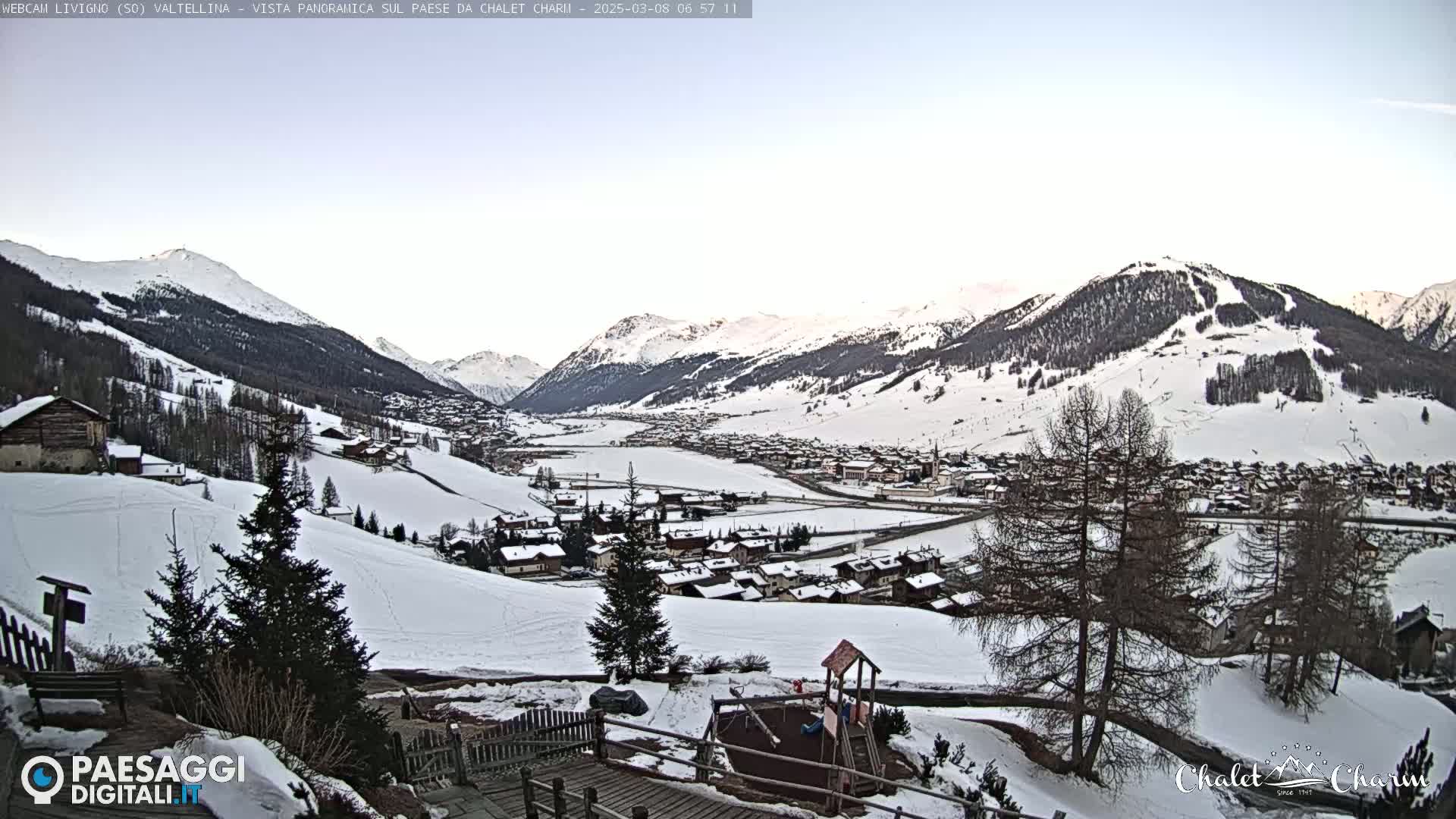 A snow-covered village nestled in a valley between snow-capped mountains under a clear, light-blue sky.