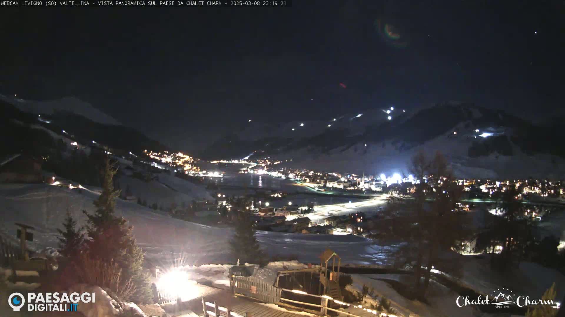 A nighttime panoramic view of a snow-covered town illuminated by lights, nestled in a valley under a clear, dark sky.