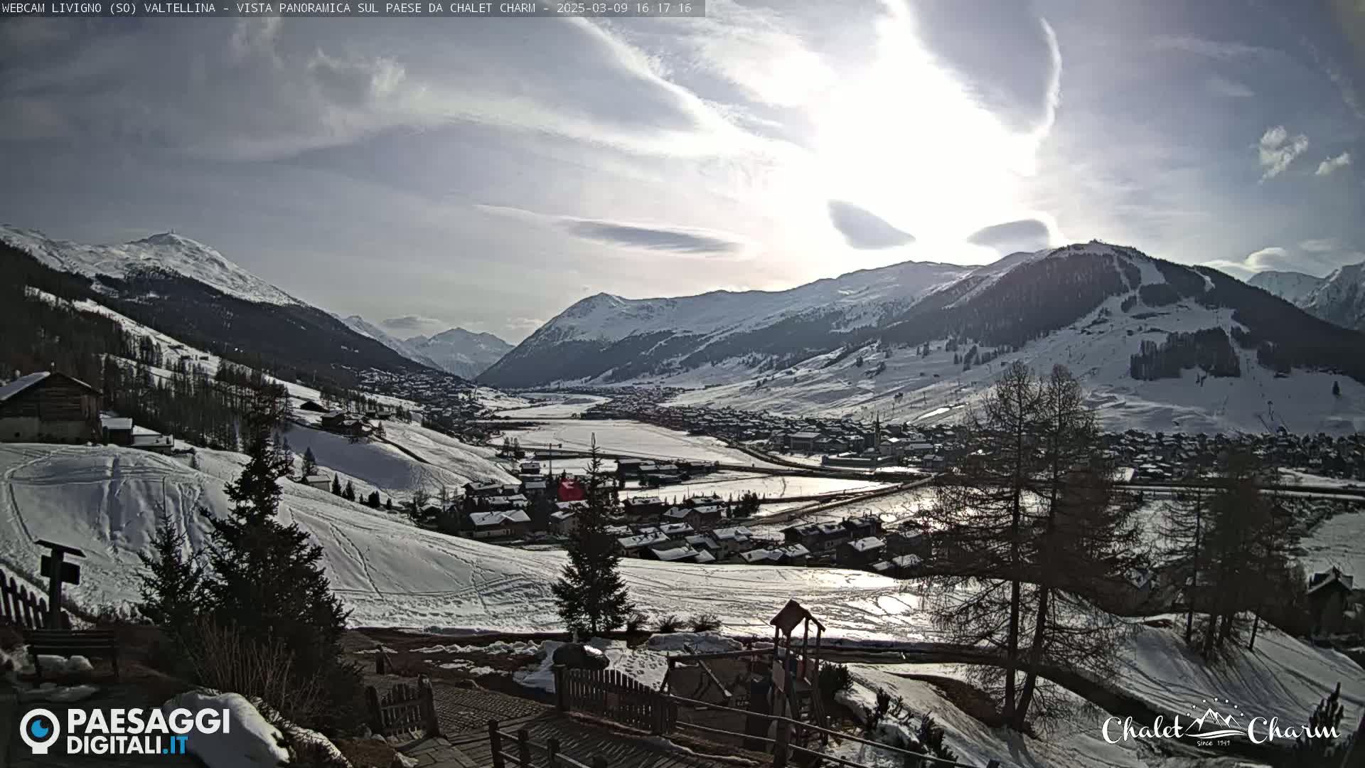 A snow-covered mountain valley town is seen on a partly sunny day, with snow-covered hills and mountains surrounding it.