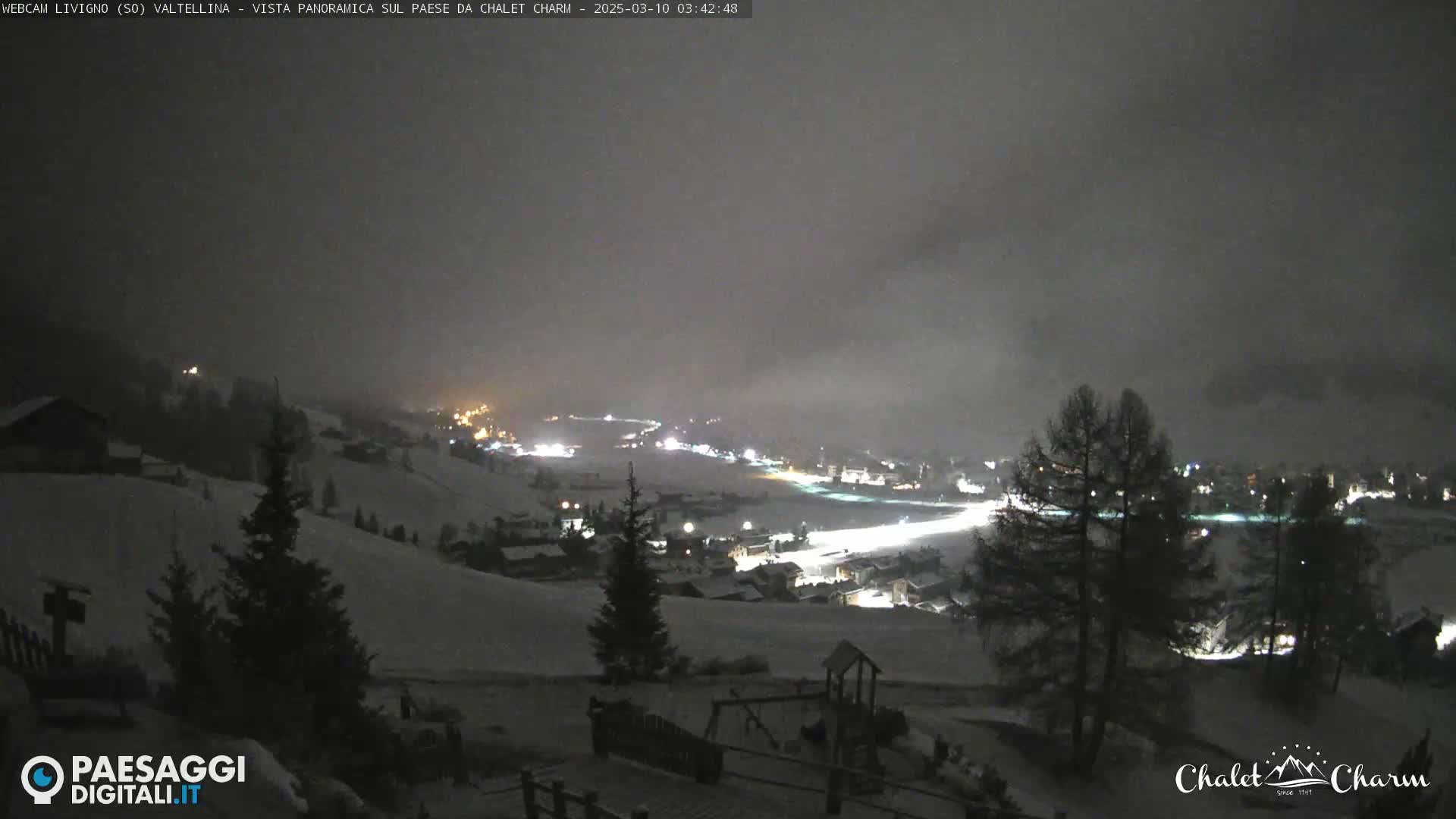 A snow-covered hillside at night, with a village illuminated below in foggy conditions.