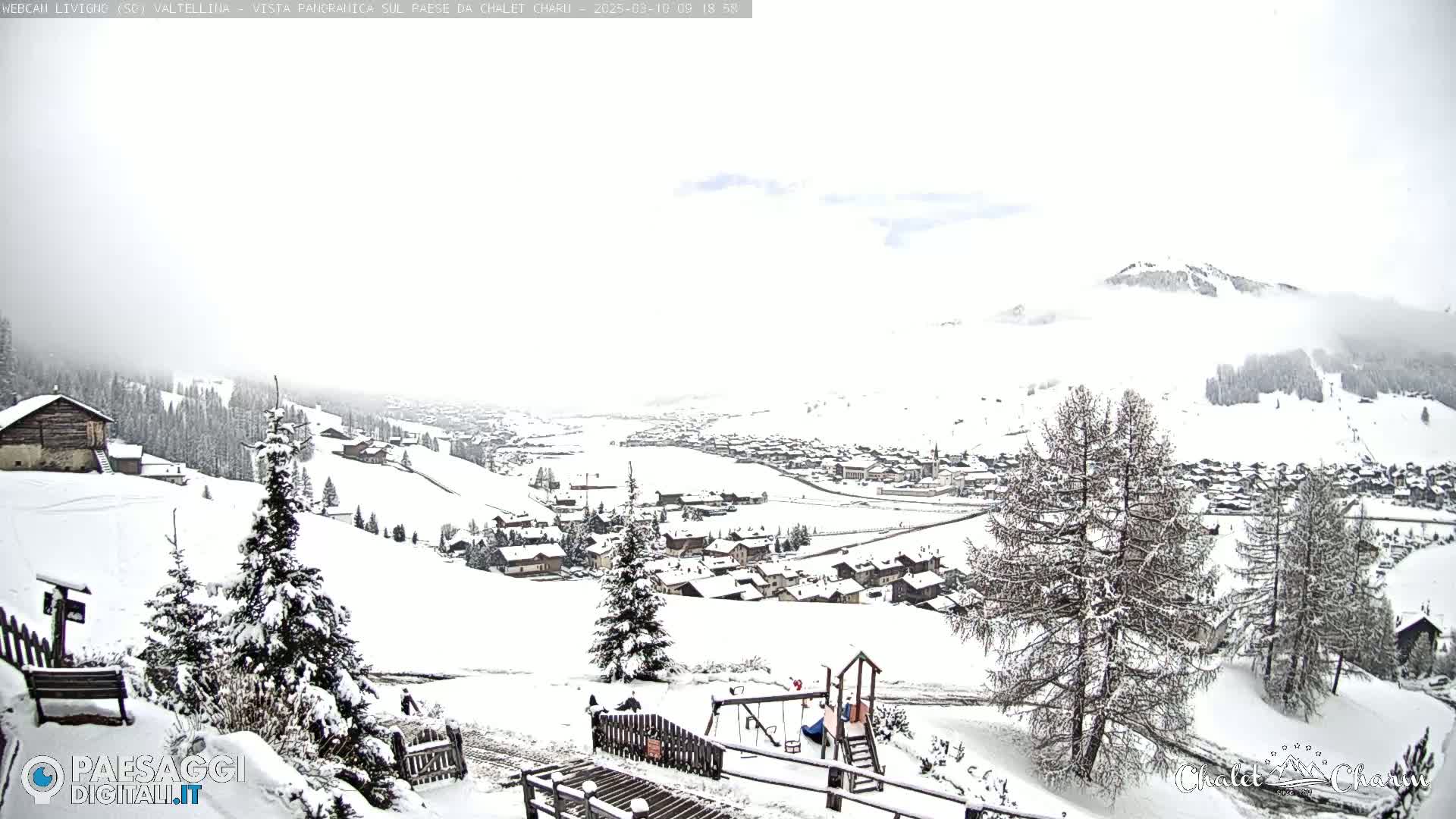 A snow-covered village nestled in a valley, with snow-laden trees and a children's playground in the foreground, under an overcast sky.