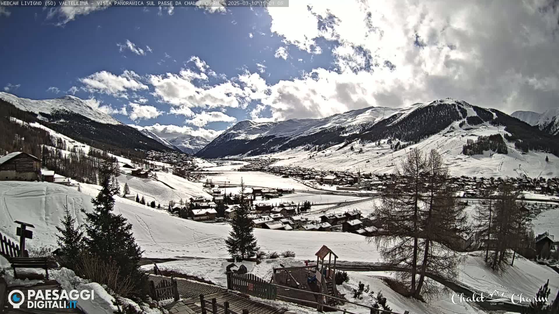 A snow-covered village nestled in a valley between snow-capped mountains under a partly cloudy sky.