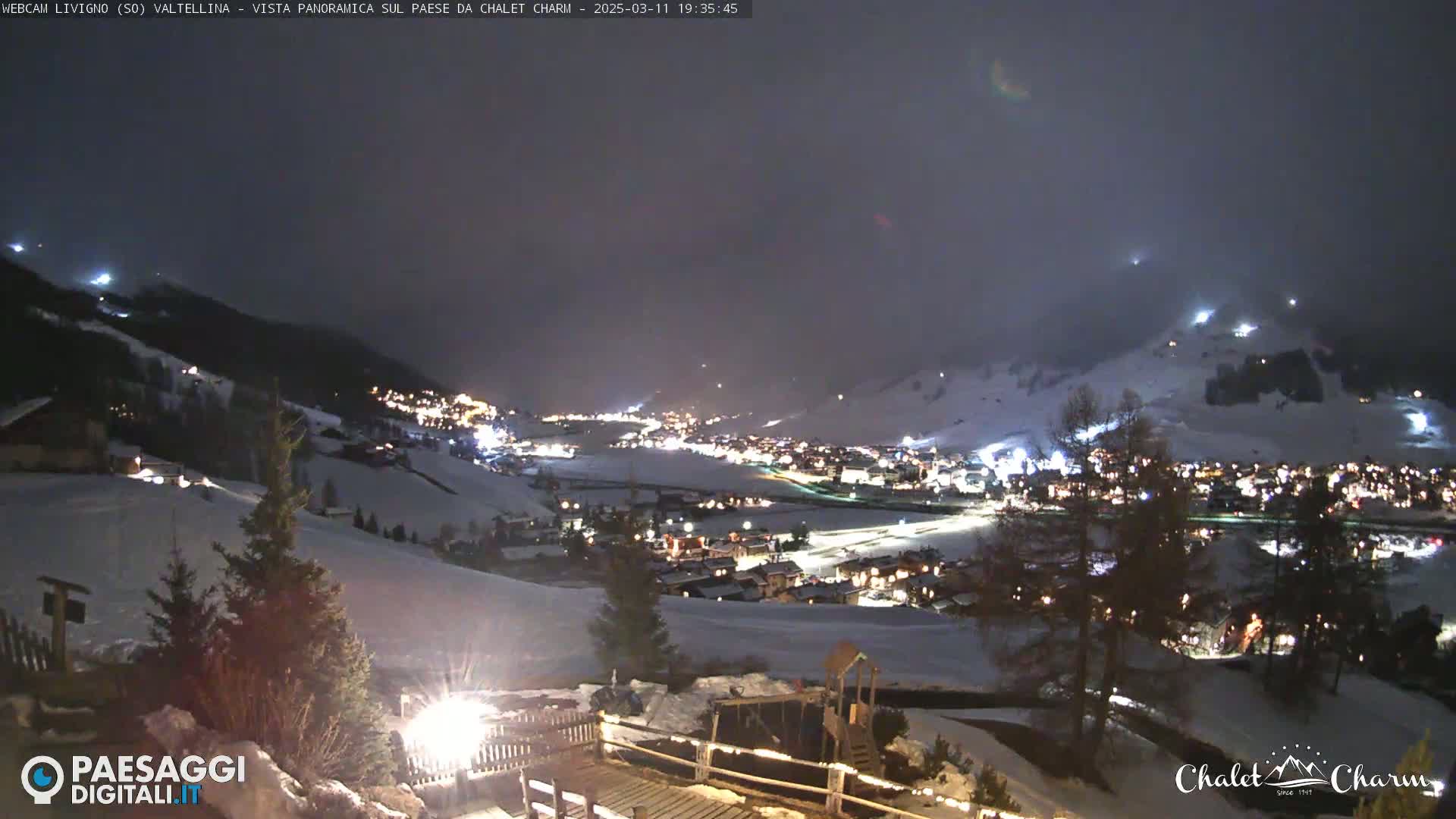 A nighttime, snowy, and hazy view of a village nestled in a valley, illuminated by lights, with a few trees and a playground in the foreground.
