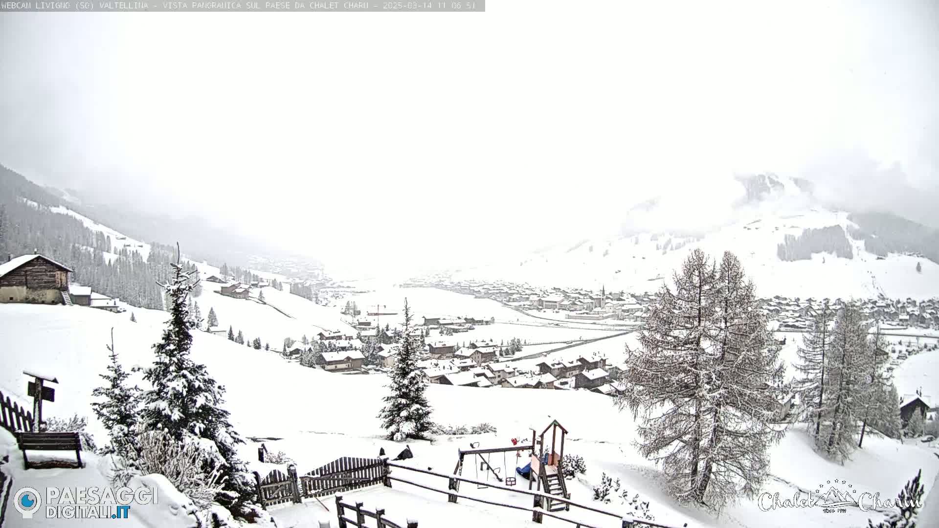A snow-covered village nestled in a valley, with snow-laden trees and a playground in the foreground, under a heavily overcast sky.