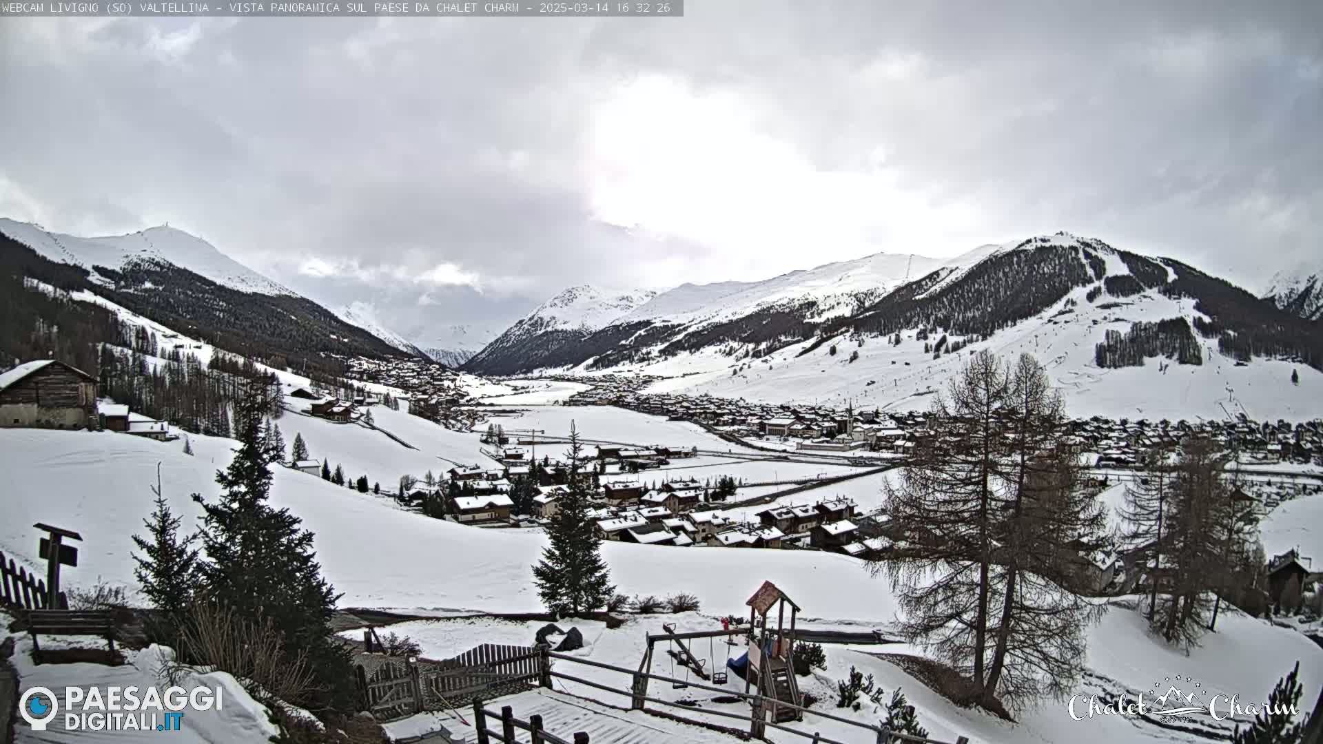 A snow-covered village nestled in a valley between snow-capped mountains under a cloudy sky.