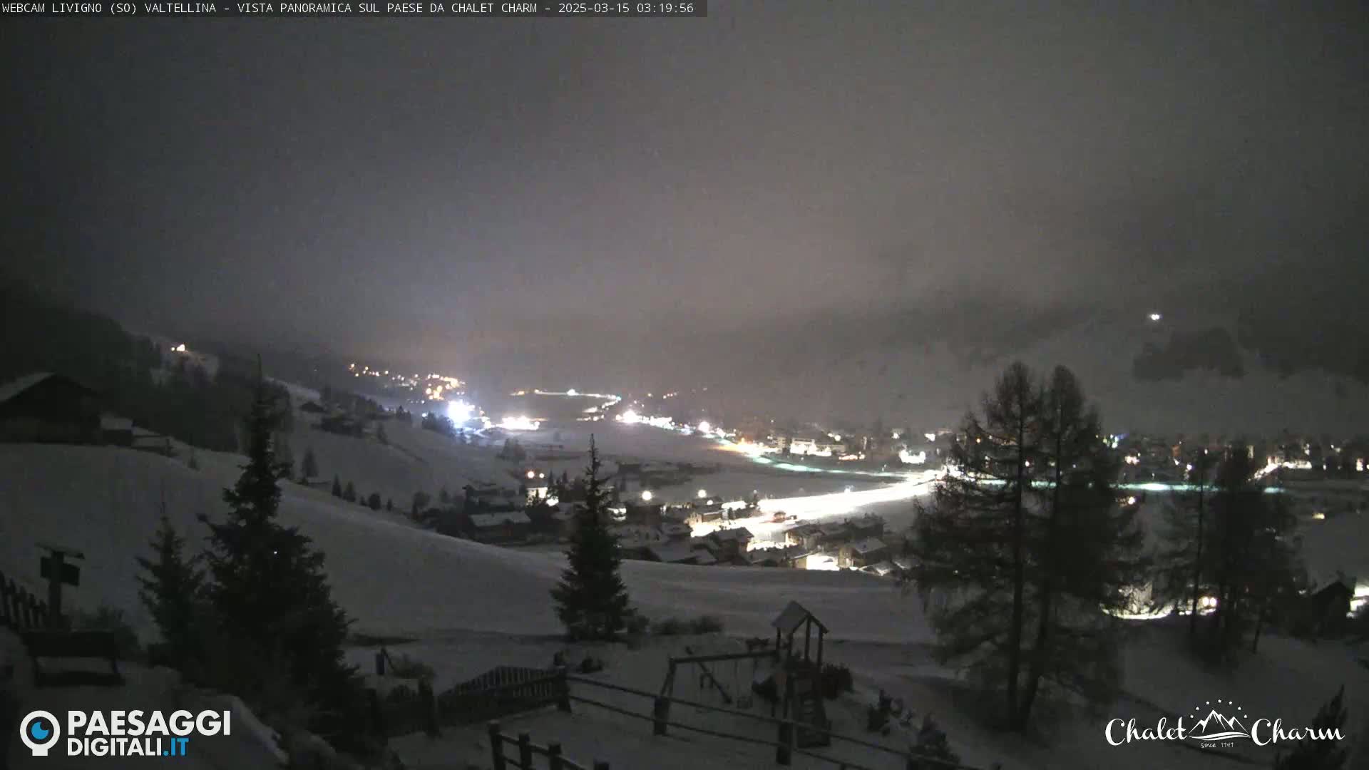 A nighttime, snowy, panoramic view of a village illuminated by streetlights under a dark, overcast sky.