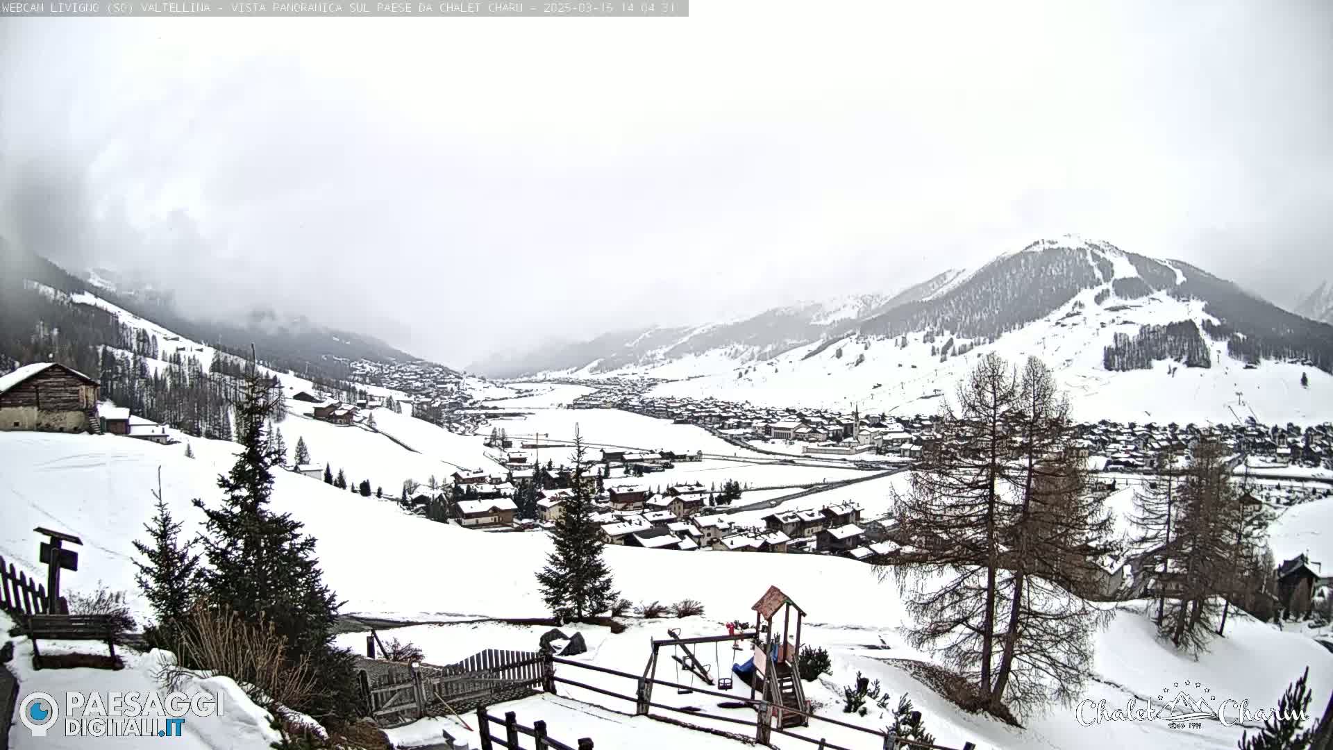 A snow-covered village nestled in a valley between snow-capped mountains under an overcast sky.