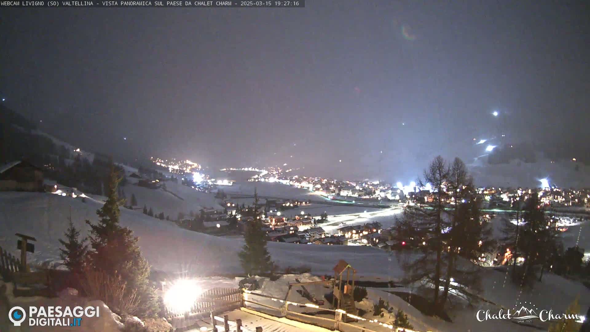 A nighttime panoramic view of a snow-covered town illuminated by lights, under a hazy, dark sky.
