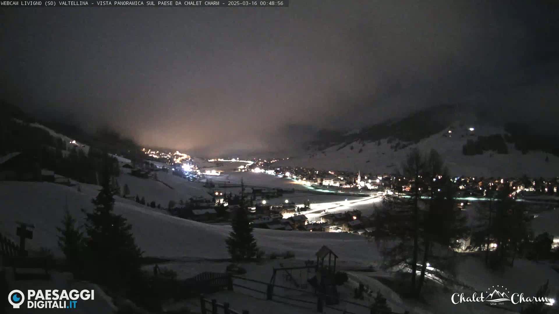 A nighttime panoramic view of a snow-covered town illuminated by lights under a hazy, dark sky.