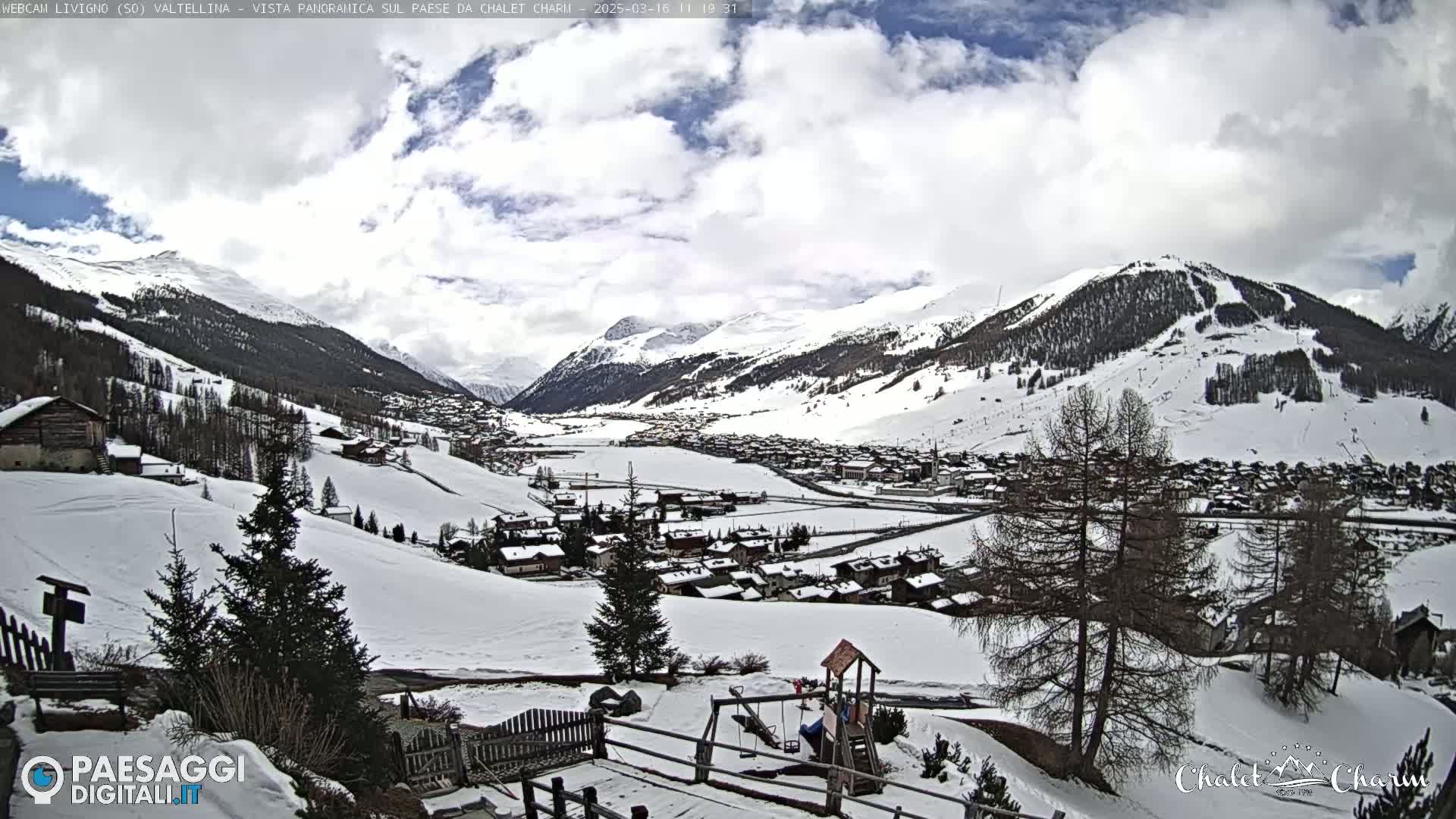 A snow-covered mountain village is seen under a partly cloudy sky, with a playground visible in the foreground.