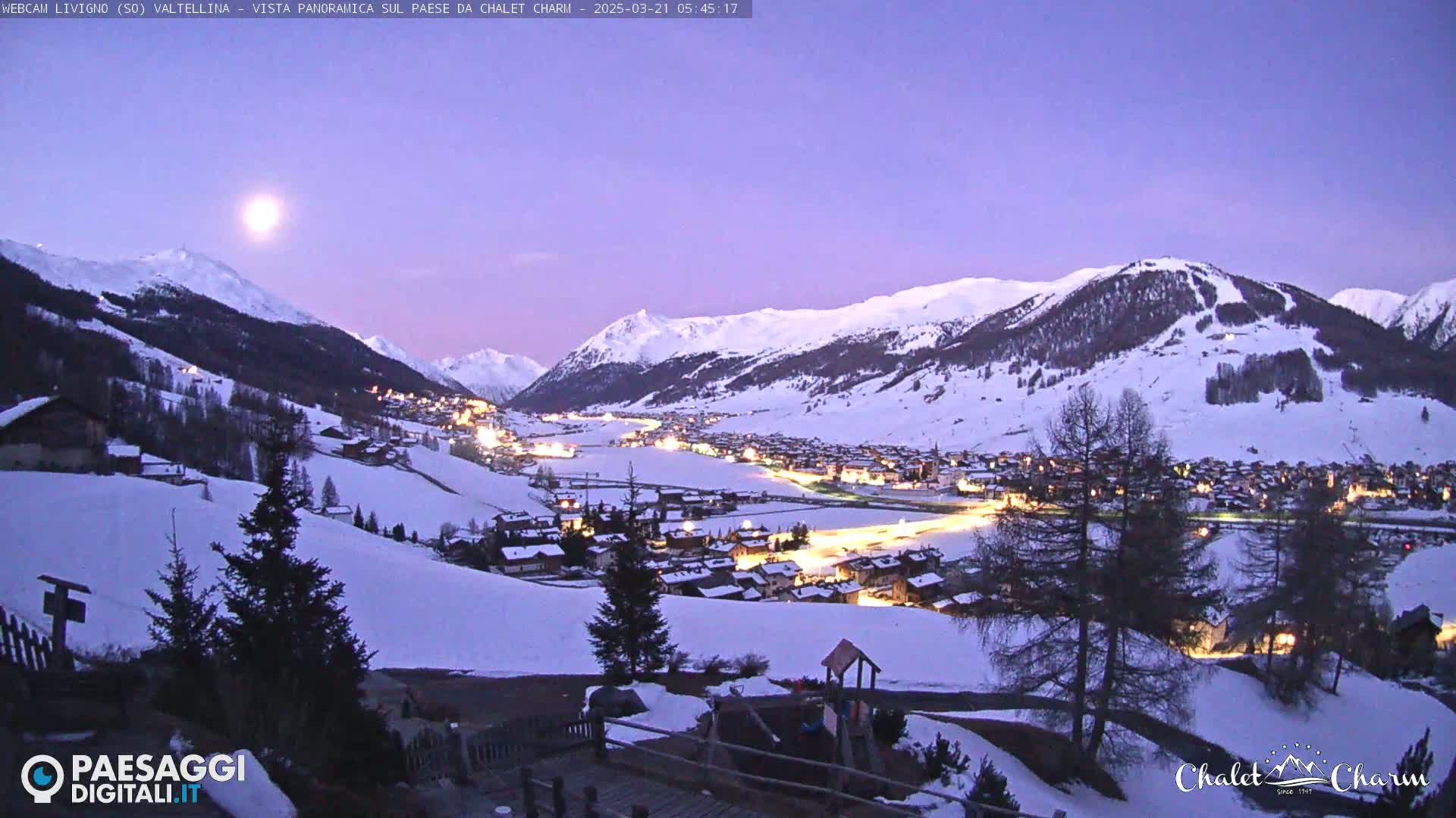 A snow-covered village nestled in a valley between snow-capped mountains is illuminated at twilight under a full moon.