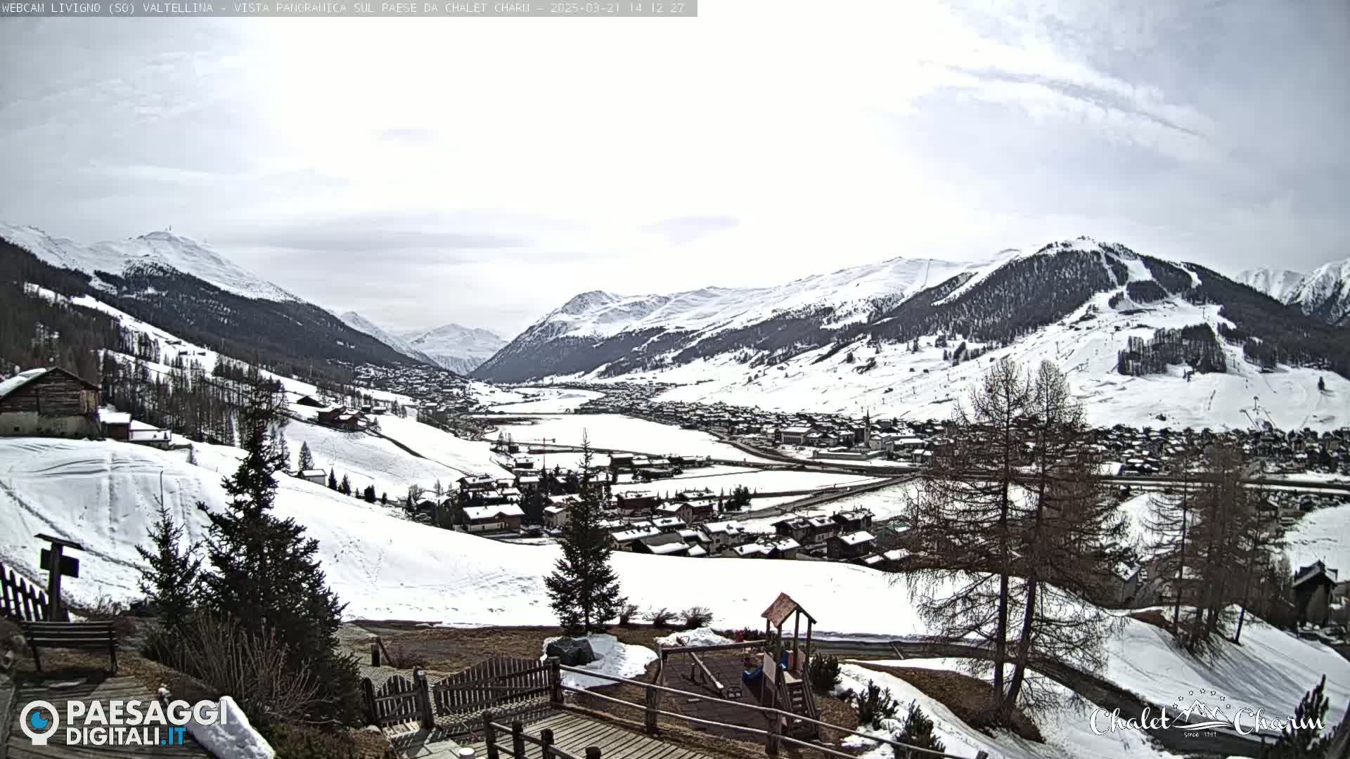 A snow-covered mountain village is nestled in a valley between snow-capped mountains under an overcast sky.