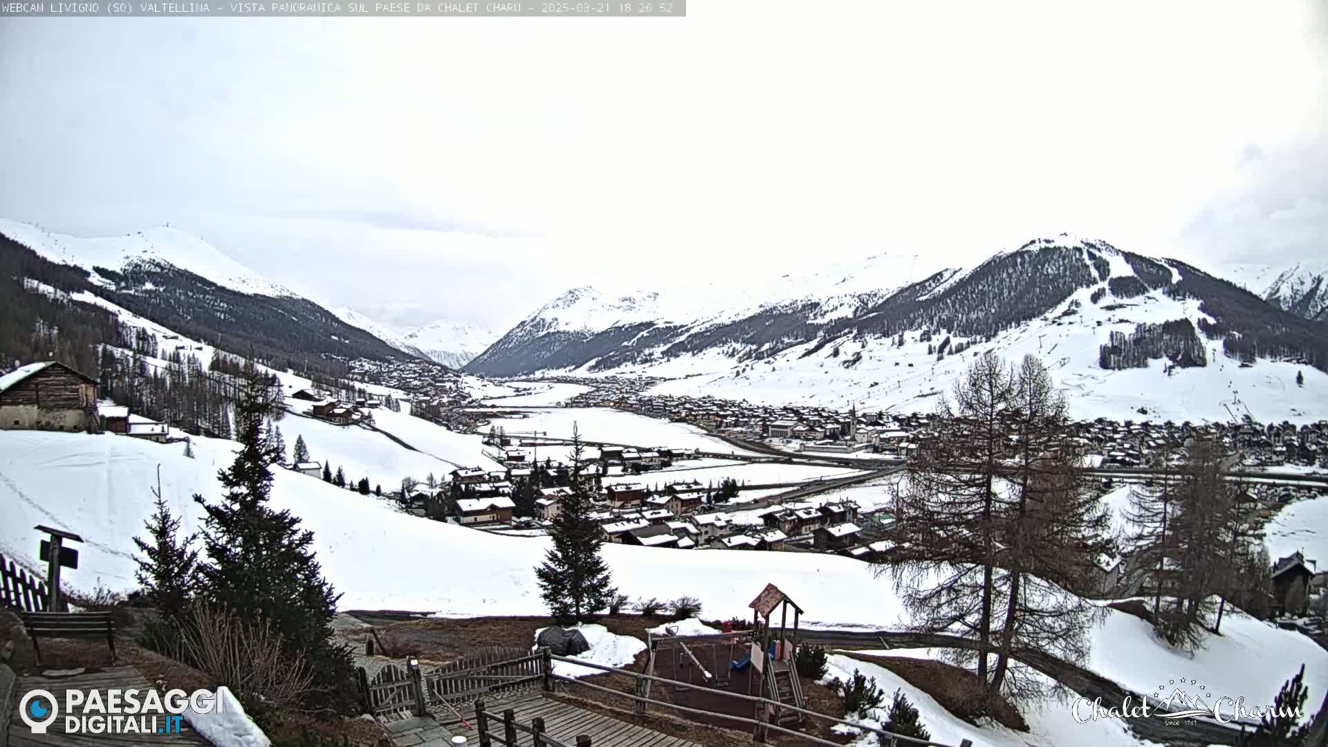 A snow-covered mountain village is nestled in a valley between snow-capped mountains under an overcast sky.