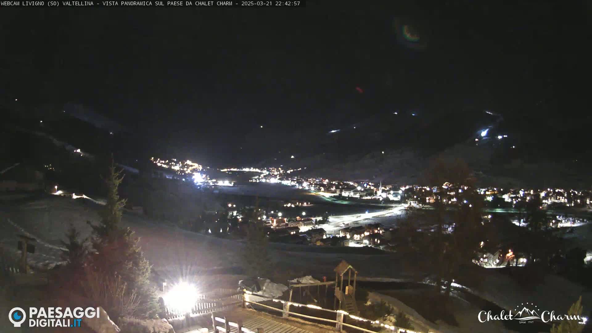 A nighttime, snow-covered landscape shows a small town illuminated below, with hills and mountains in the background under a clear, dark sky.