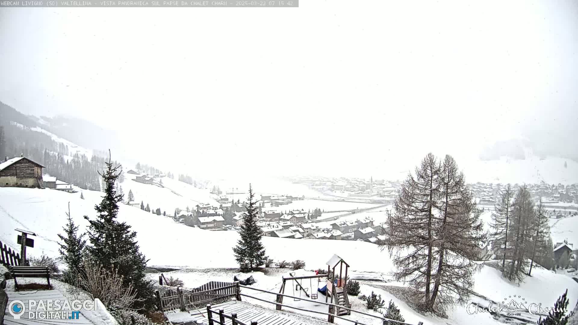 A snow-covered village is seen from a hillside vantage point, with light snowfall and a playground in the foreground.