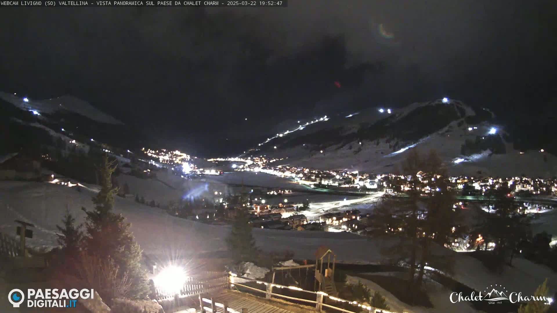 A nighttime, panoramic view of a snow-covered town nestled in a valley, illuminated by lights, with ski slopes visible on surrounding hills.