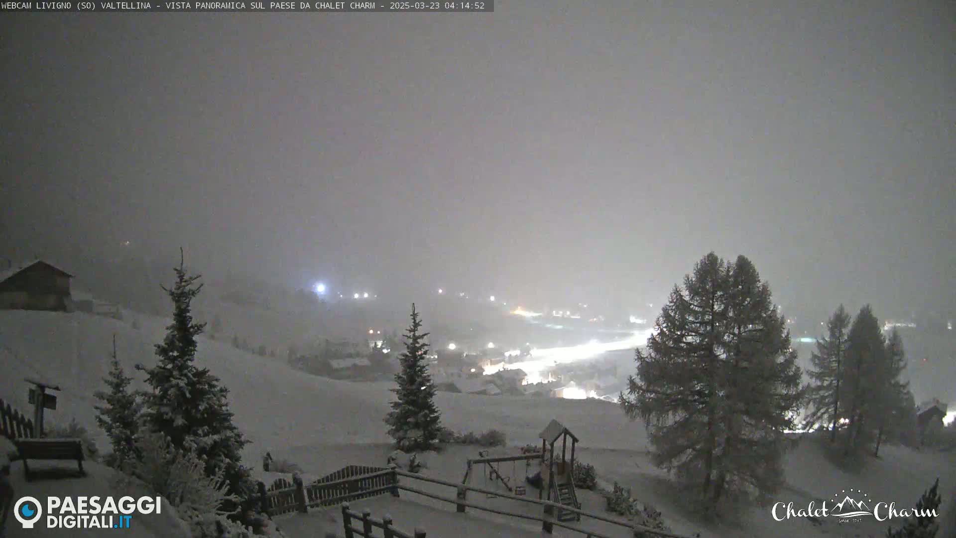 A snow-covered landscape at night, with a village illuminated in the distance under a hazy, dark sky.