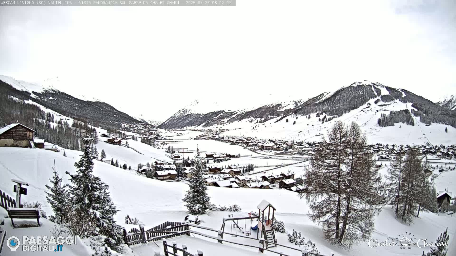A snow-covered village nestled in a valley between snow-capped mountains under an overcast sky.