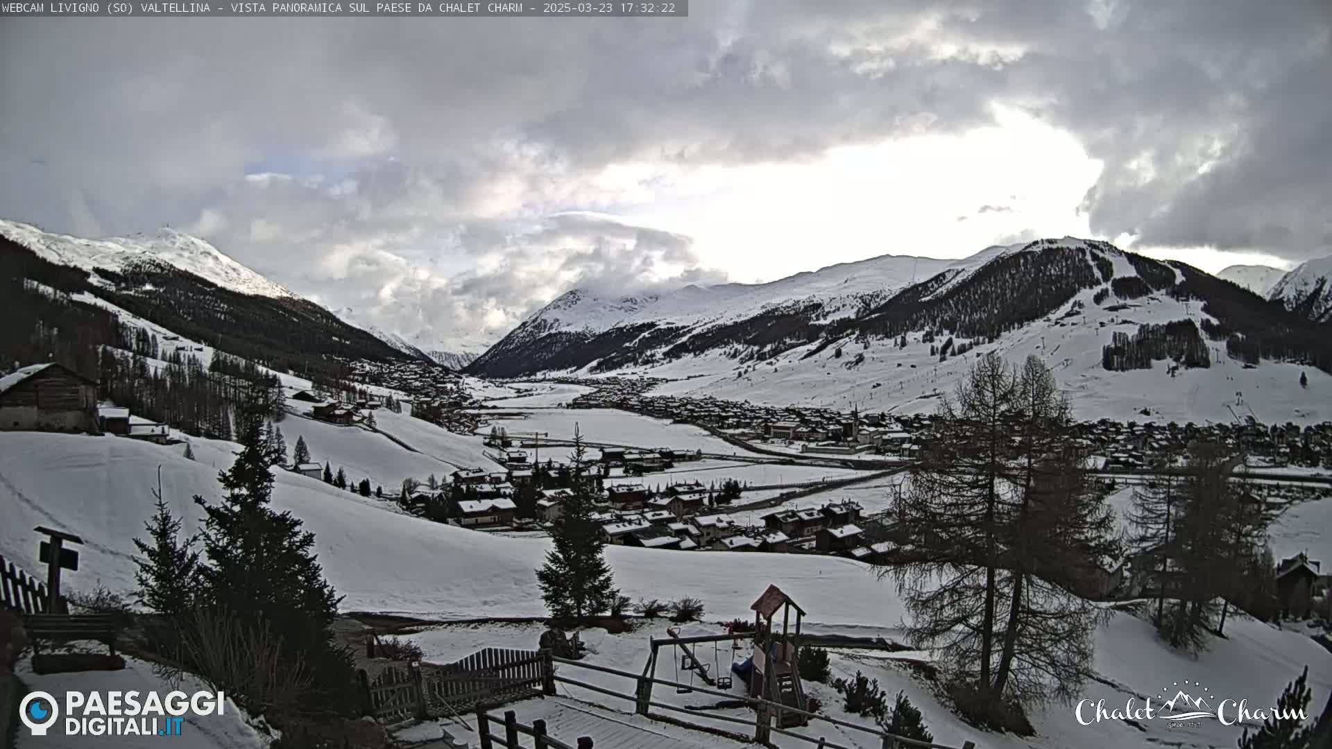 A snow-covered village nestled in a valley between snow-capped mountains under a cloudy sky.