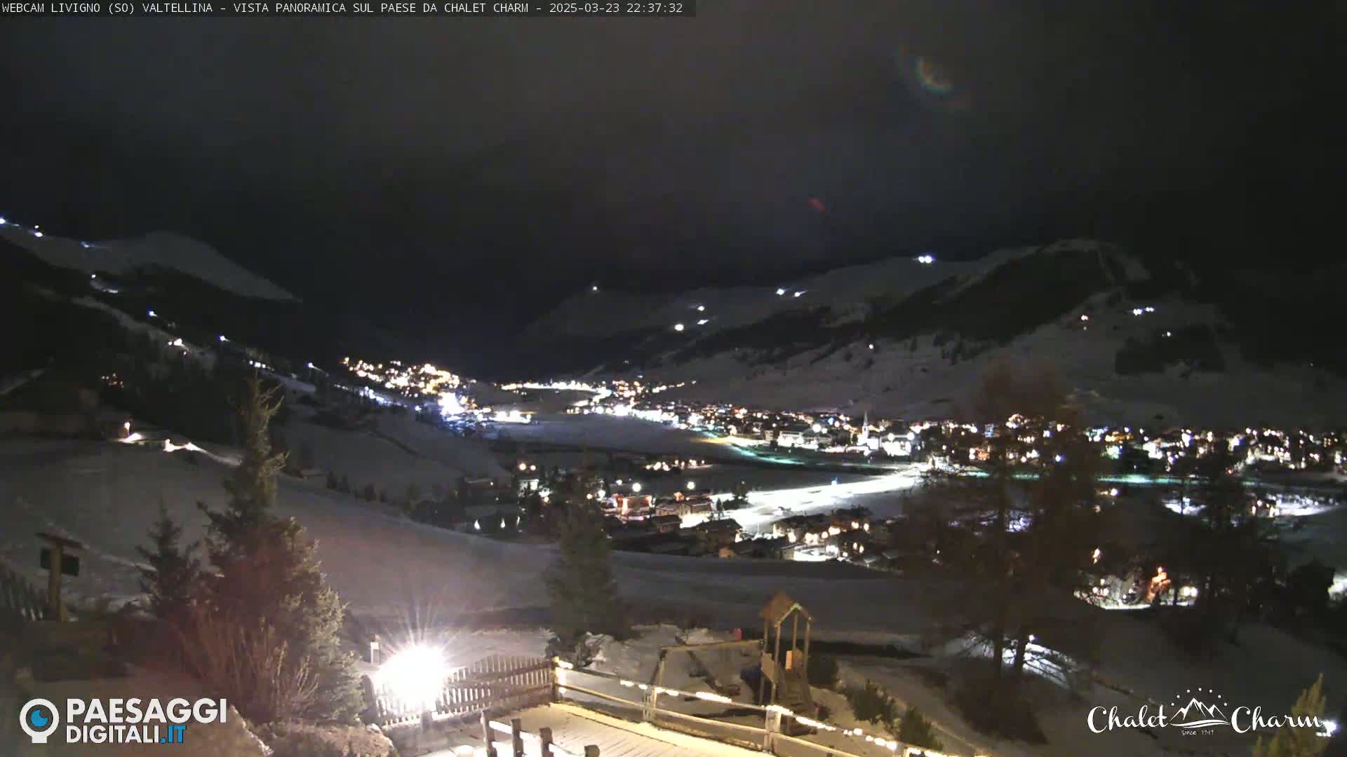 A nighttime panoramic view of a snow-covered town nestled in a valley, illuminated by lights, under a dark sky.