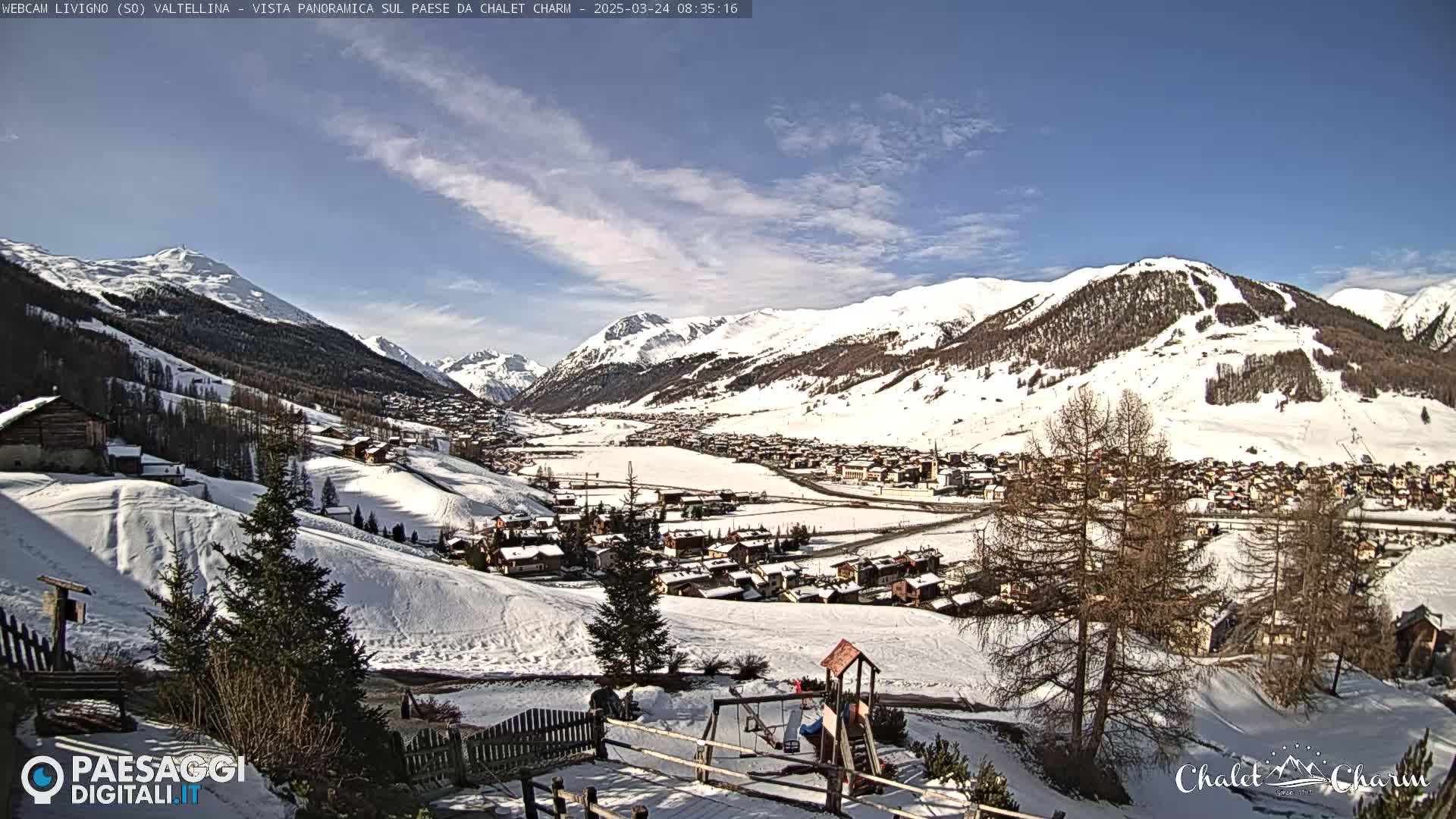 A snow-covered village nestled in a valley between snow-capped mountains under a mostly sunny sky.